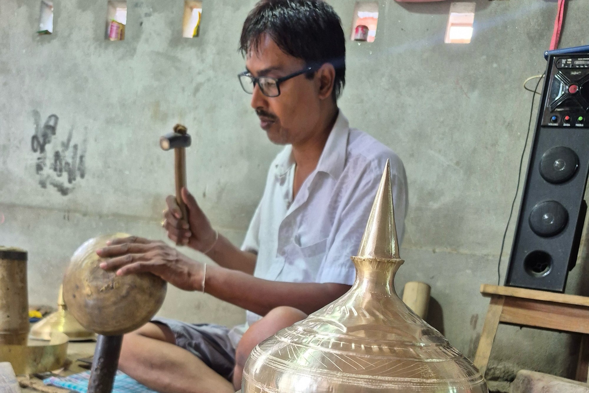 Haridas Das seated cross-legged on the workshop floor in glasses and a white shirt, hammering a rounded metal dome held over an iron stake, the polished lid and spire of a finished xorai in sharp focus in front of him.