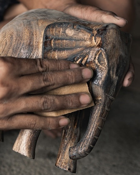 Two hands smoothing a carved wooden elephant with a fold of sandpaper, the grain of the wood showing through its dark finish.