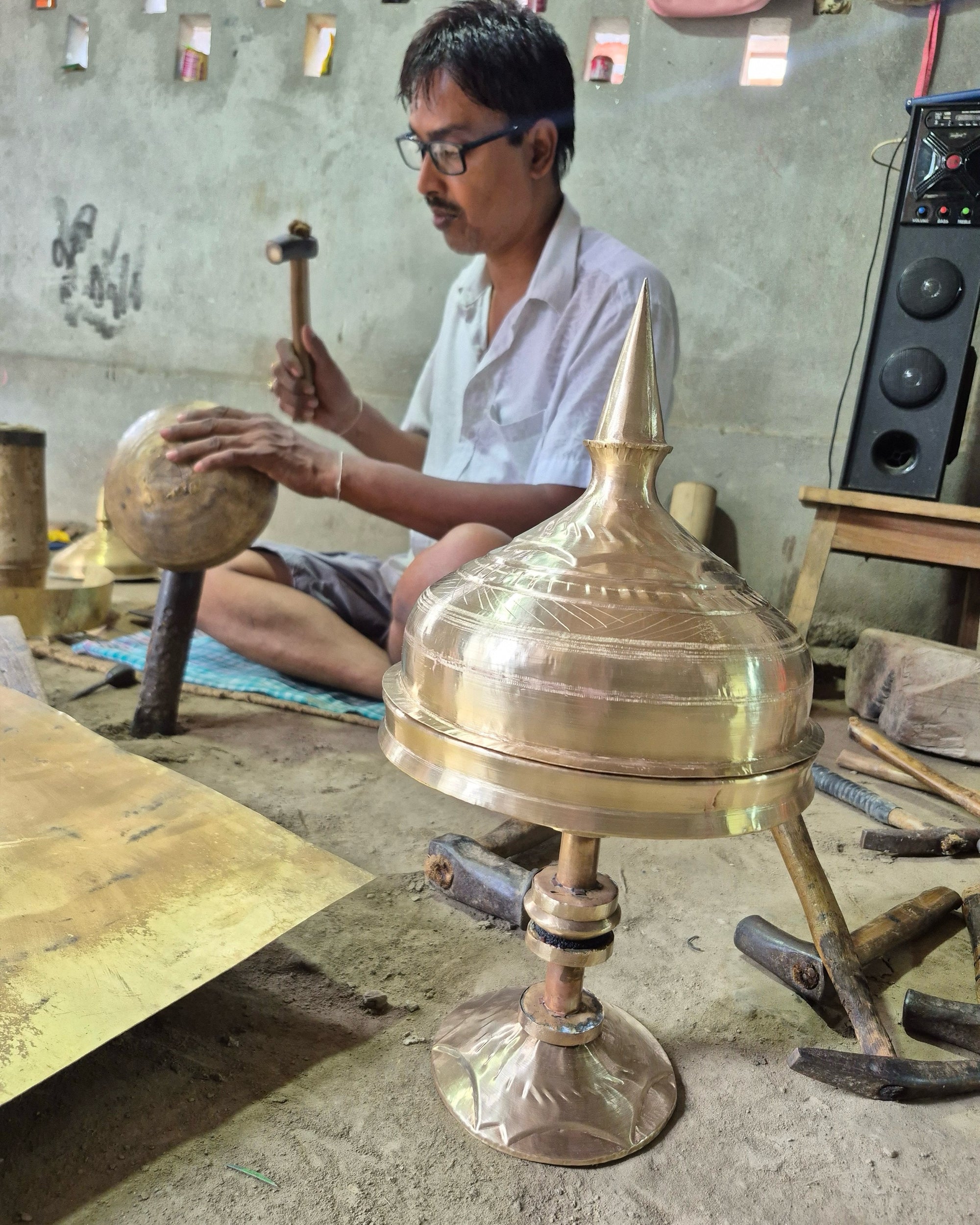 Haridas Das seated cross-legged on the workshop floor, hammering a rounded brass dome over an iron stake, the lid and spire of a finished xorai in front of him.