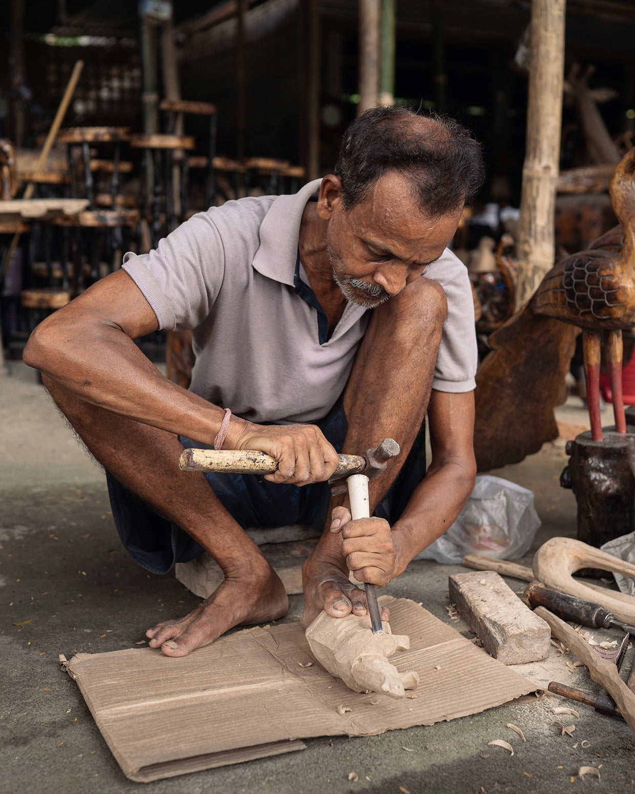 A carver squatting on a workshop floor, a chisel in one hand and a hammer raised in the other, over a pale block of wood held down by his foot.