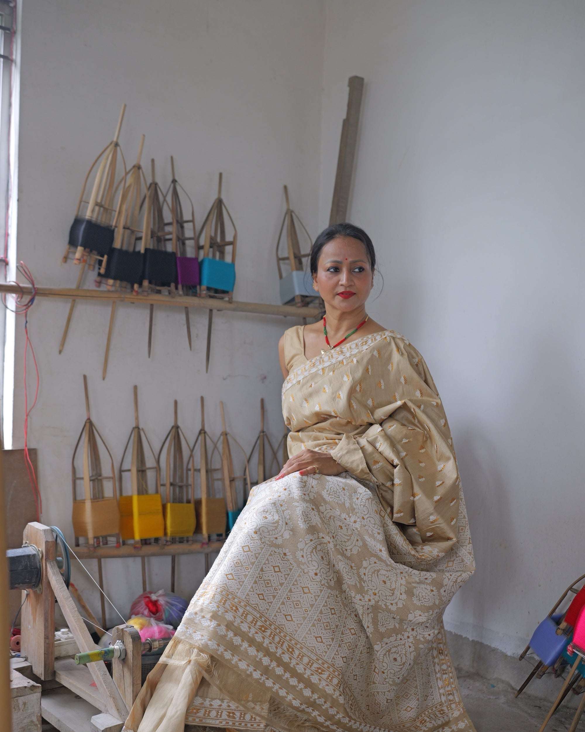 Seated beside a wall of bamboo reels wound with coloured yarn, a wooden winding frame at her side. The saree crosses the shoulder and falls across the lap to the floor: a soft gold body with small ivory motifs above a lower fall densely worked in ivory.