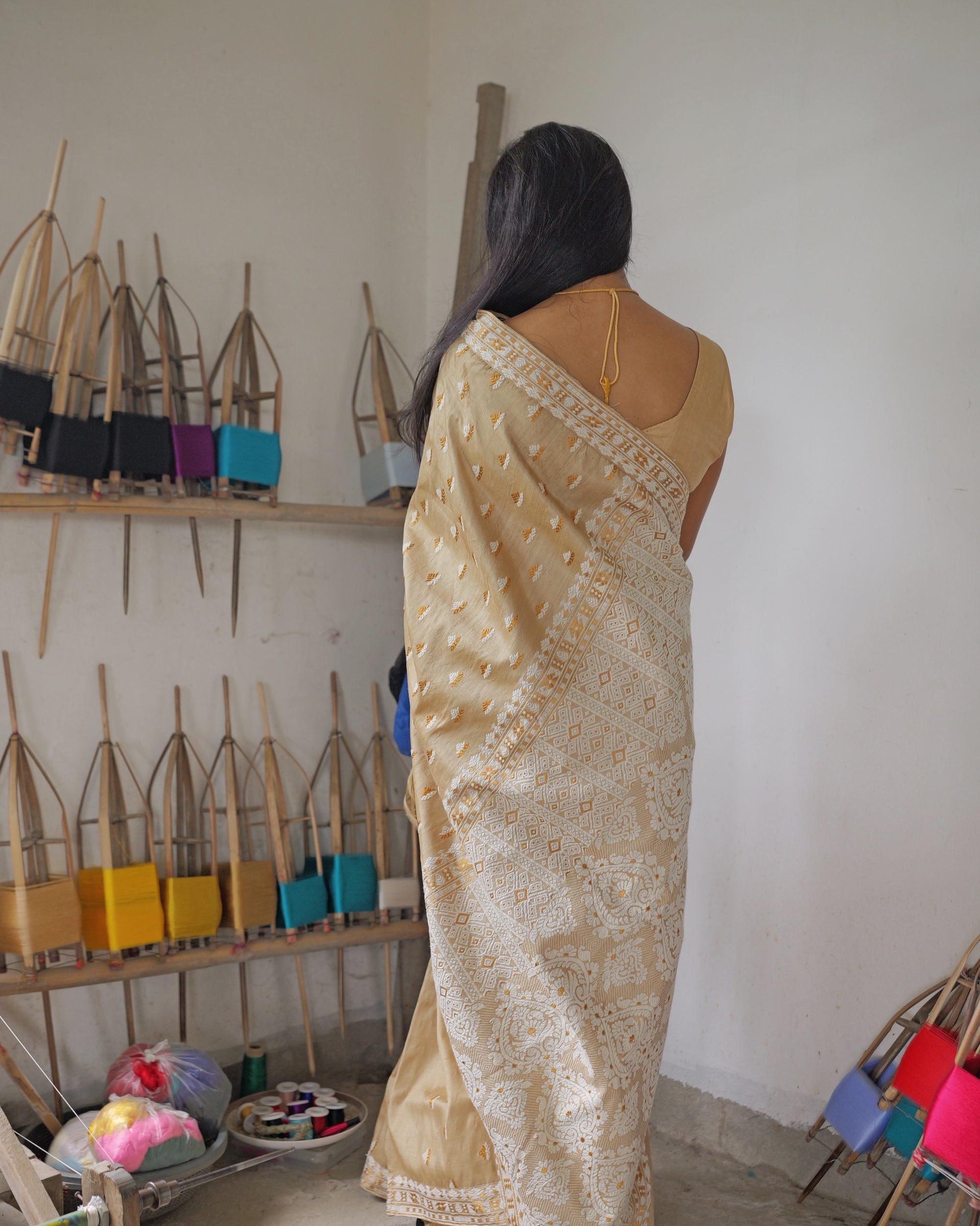 Seen from behind, standing by the wall of bamboo reels, the saree falling full length to the floor and the worked pallu hanging down the back.