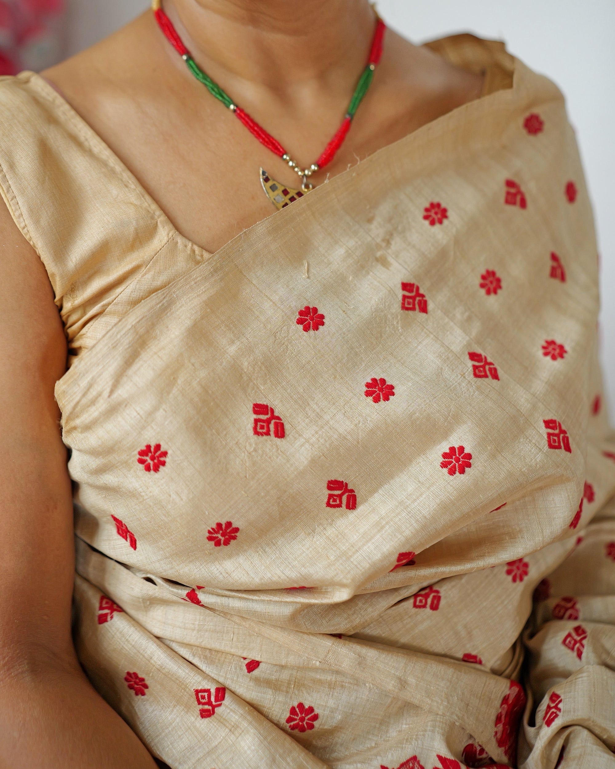 The drape across the chest, close: small red eight-petal flowers and stepped blocks in turn on gold silk, below a red and green bead necklace with a small gold pendant.