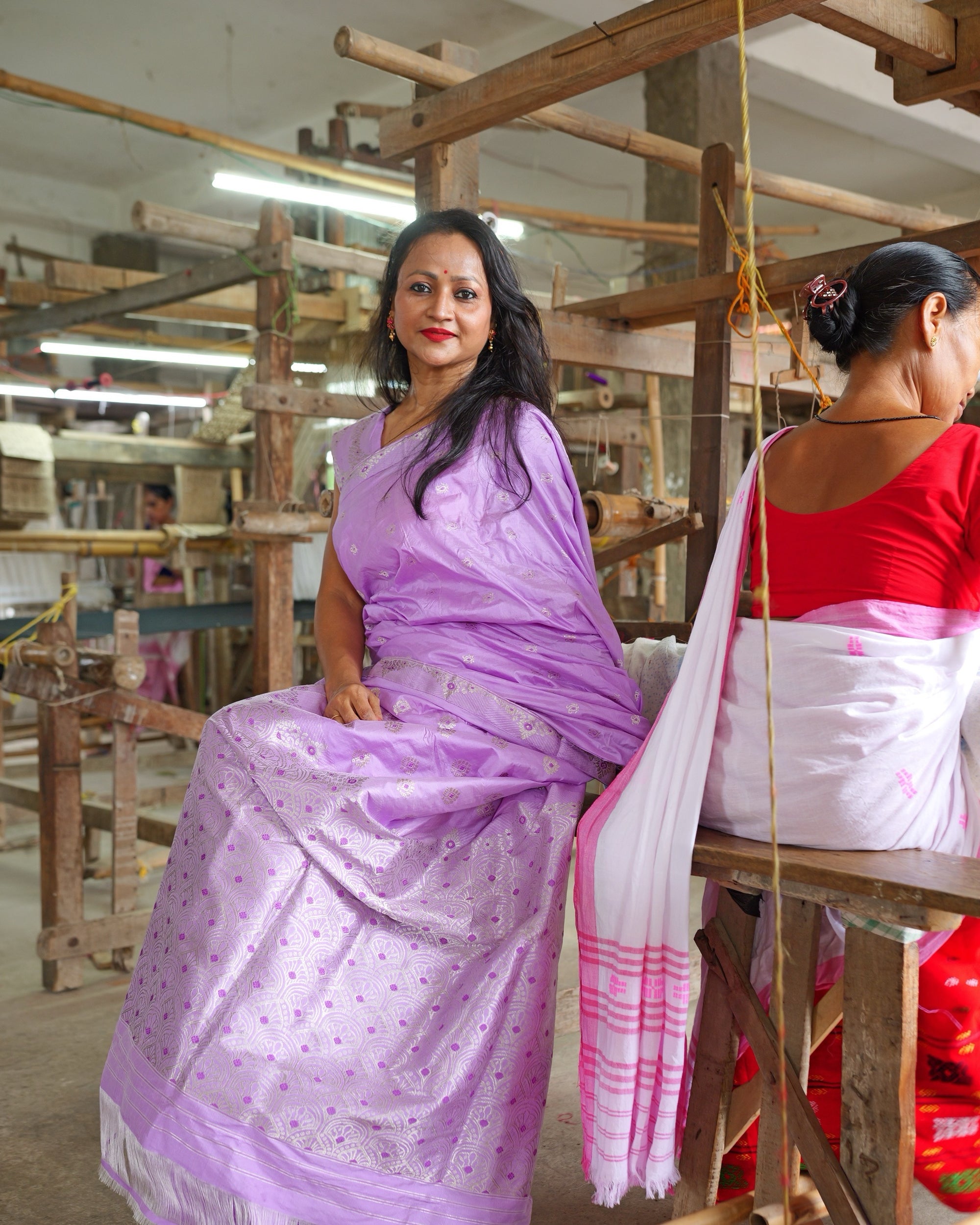 Seated at the edge of a long loom shed, with another weaver working at her loom in the same frame.