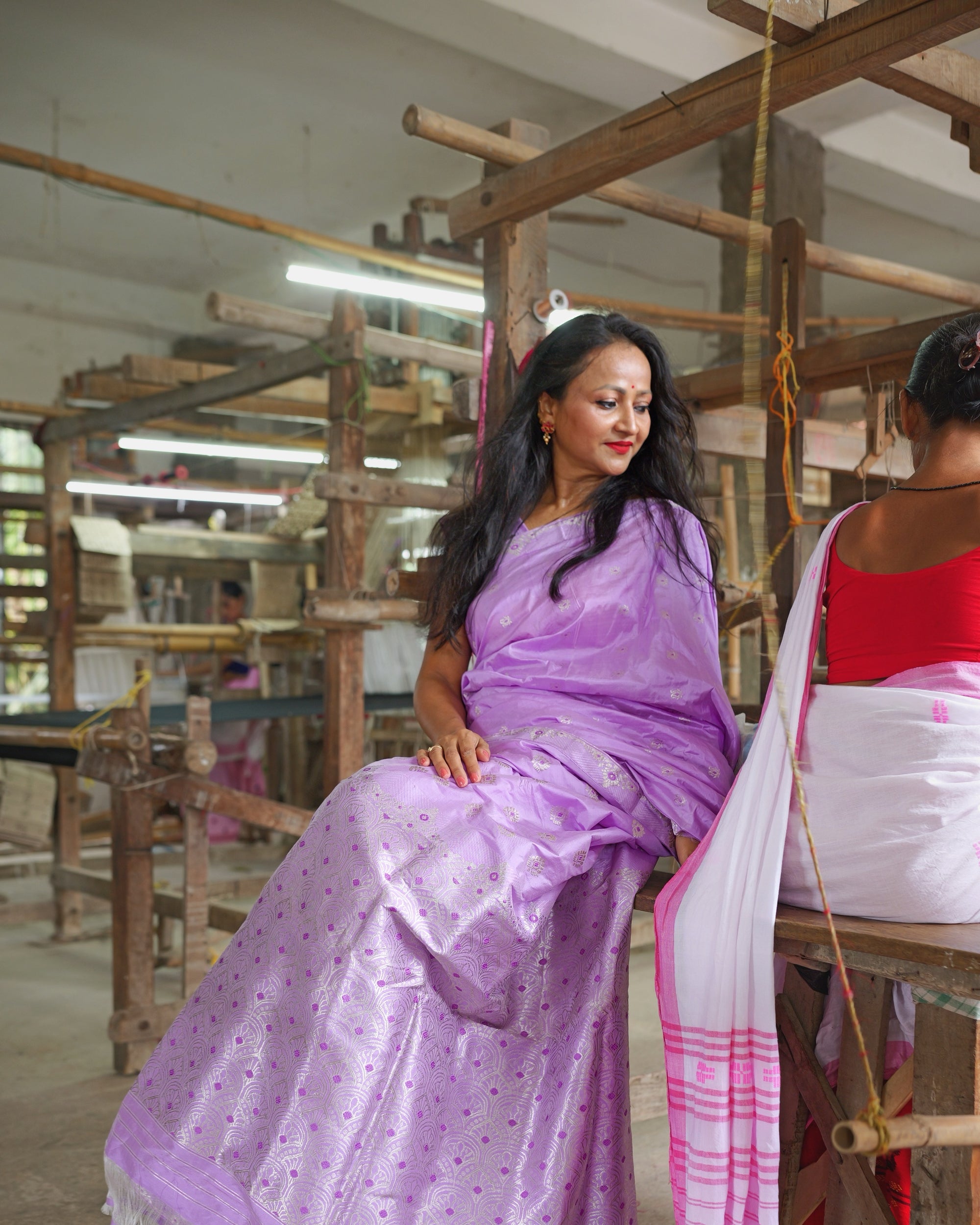 Seated in a loom shed, the whole saree visible. A pale lavender ground with a broad band of silver work along the lower drape.