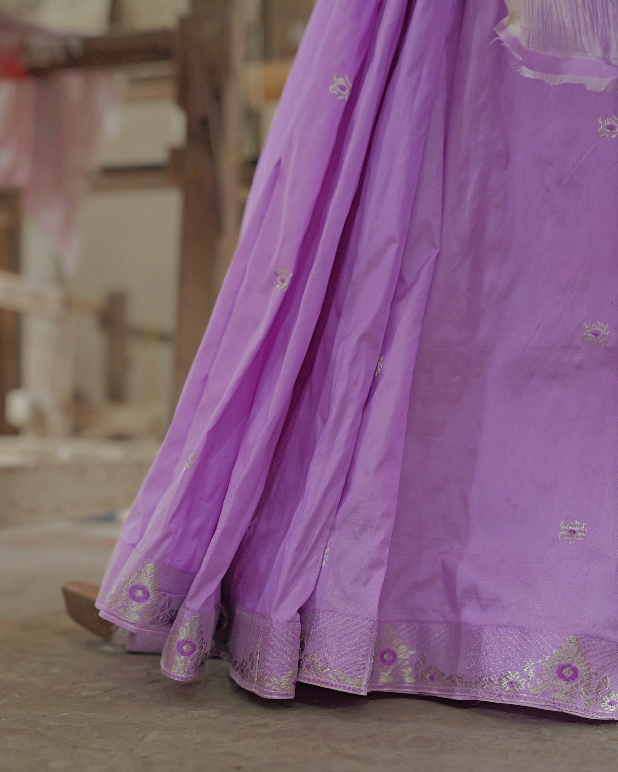 The pleats falling to a concrete floor, a silver zari border of diagonal stripes and violet-centred flowers along the hem, and in the top corner the end of the pallu: loose silver-white threads above a narrow lavender strip.