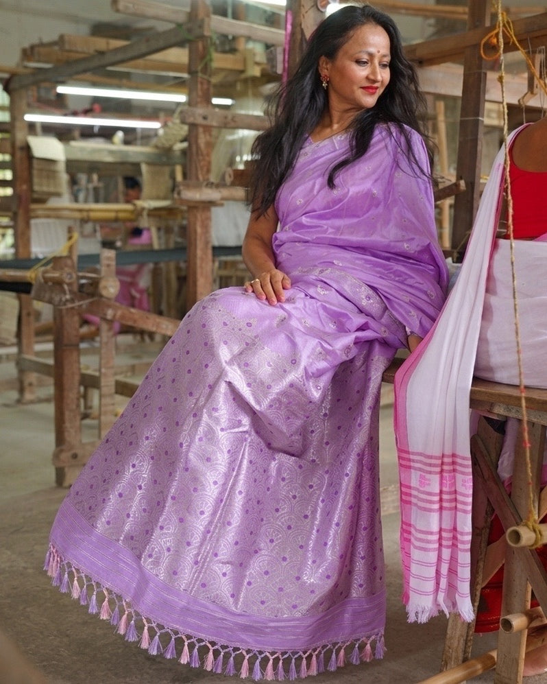 Seated on a loom bench in a weaving shed, the whole saree visible: a lilac body scattered with small violet motifs, the pallu over the shoulder and a tasselled hem, a white drape hanging beside her.