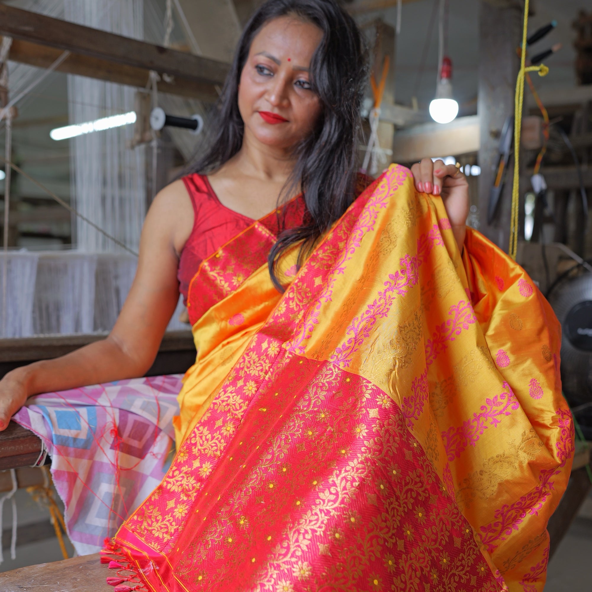 Seated against the beam of a loom, lifting the pallu at her shoulder so it falls open in front of her: a marigold body scattered with small pink kalka above a rani-pink field of gold creeper ending in pink tassels. Worn with a red sleeveless blouse, warp threads and a lamp in the shed behind.