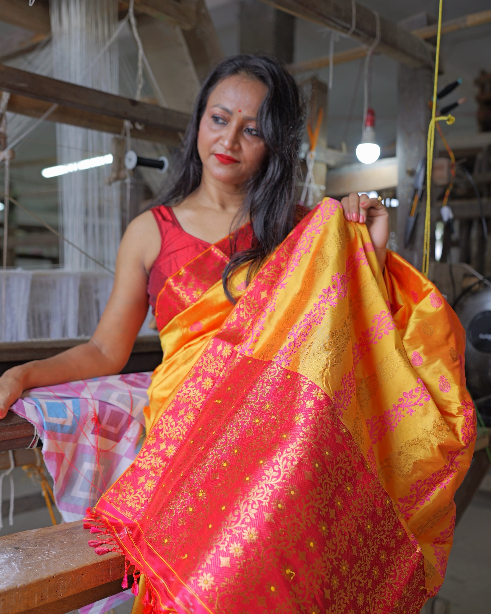 Seated against the beam of a loom, lifting the pallu at her shoulder so it falls open in front of her: a marigold body scattered with small pink kalka above a rani-pink field of gold creeper ending in pink tassels. Worn with a red sleeveless blouse, warp threads and a lamp in the shed behind.