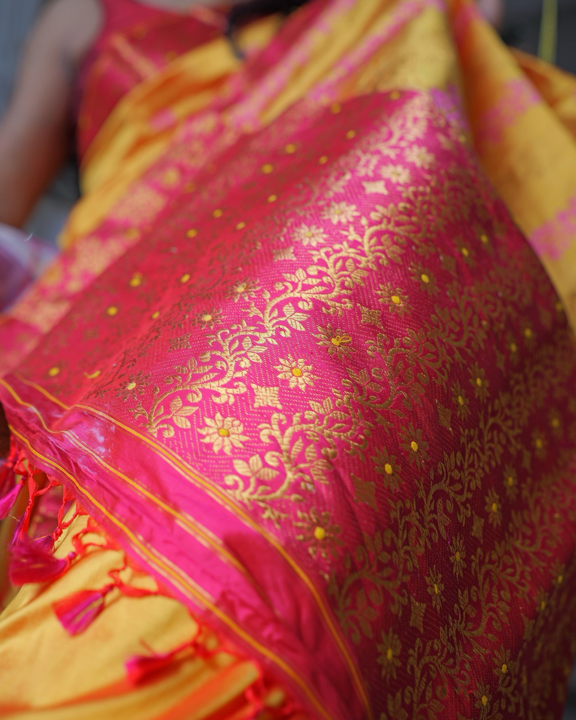 The pallu close up, held towards the camera: gold creeper and eight-petal gold flowers with yellow centres on a deep pink ground, fine yellow stripes along its end and pink tassels below.
