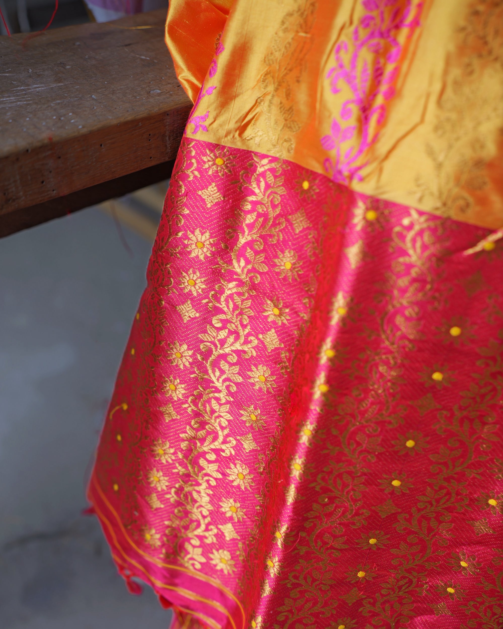 The pallu hanging over a wooden beam, where the marigold body, crossed by a pink creeper band and faint gold sprays, gives way to the pink field of gold creeper.