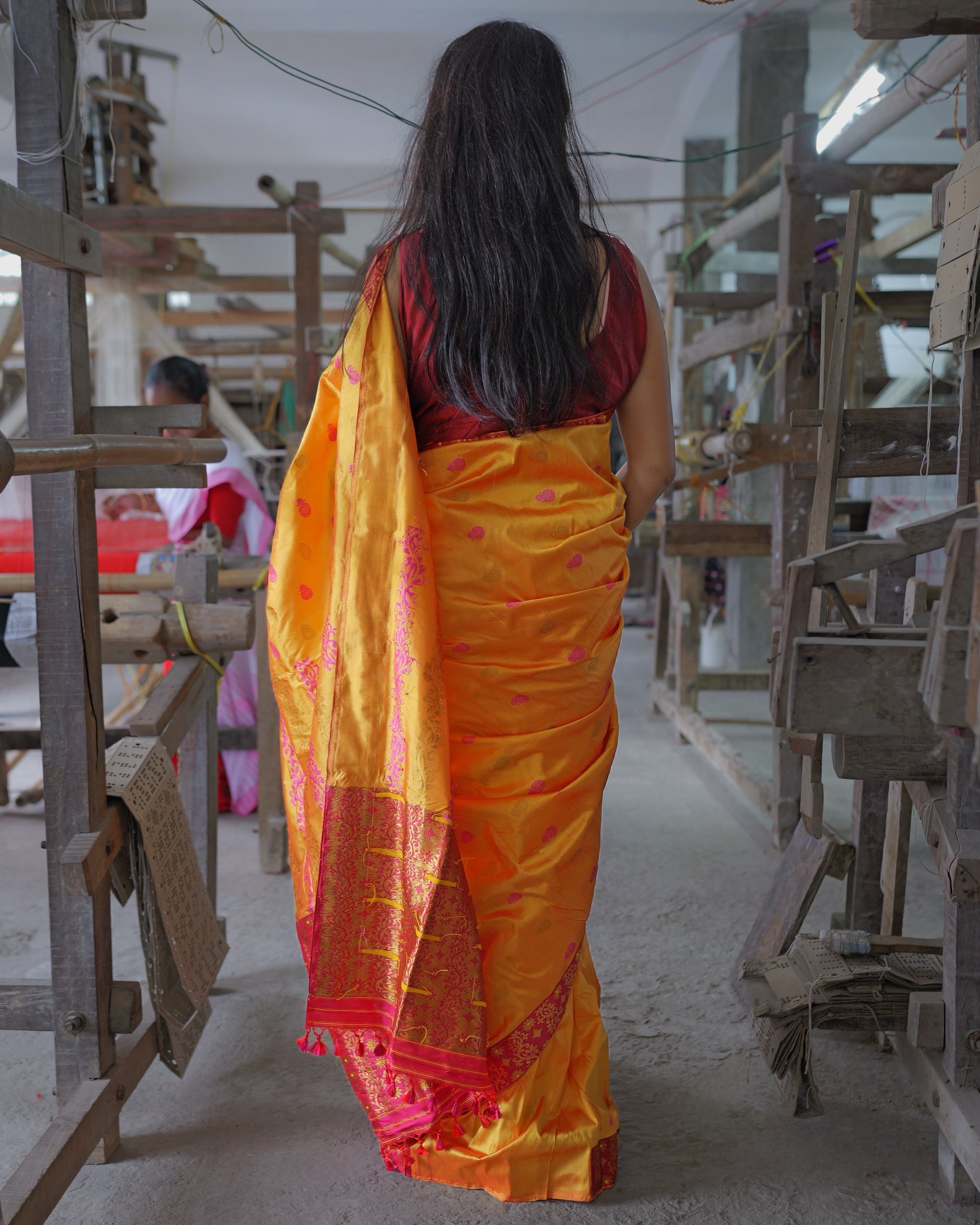 Seen from behind, full length, in an aisle between wooden looms: the marigold body dotted with small pink kalka, and the pallu falling down the back to pink tassels with its reverse turned out, mostly gold, short lengths of yellow yarn across it. A chain of punched cards hangs from the loom on the left, and another woman sits at a loom behind it.