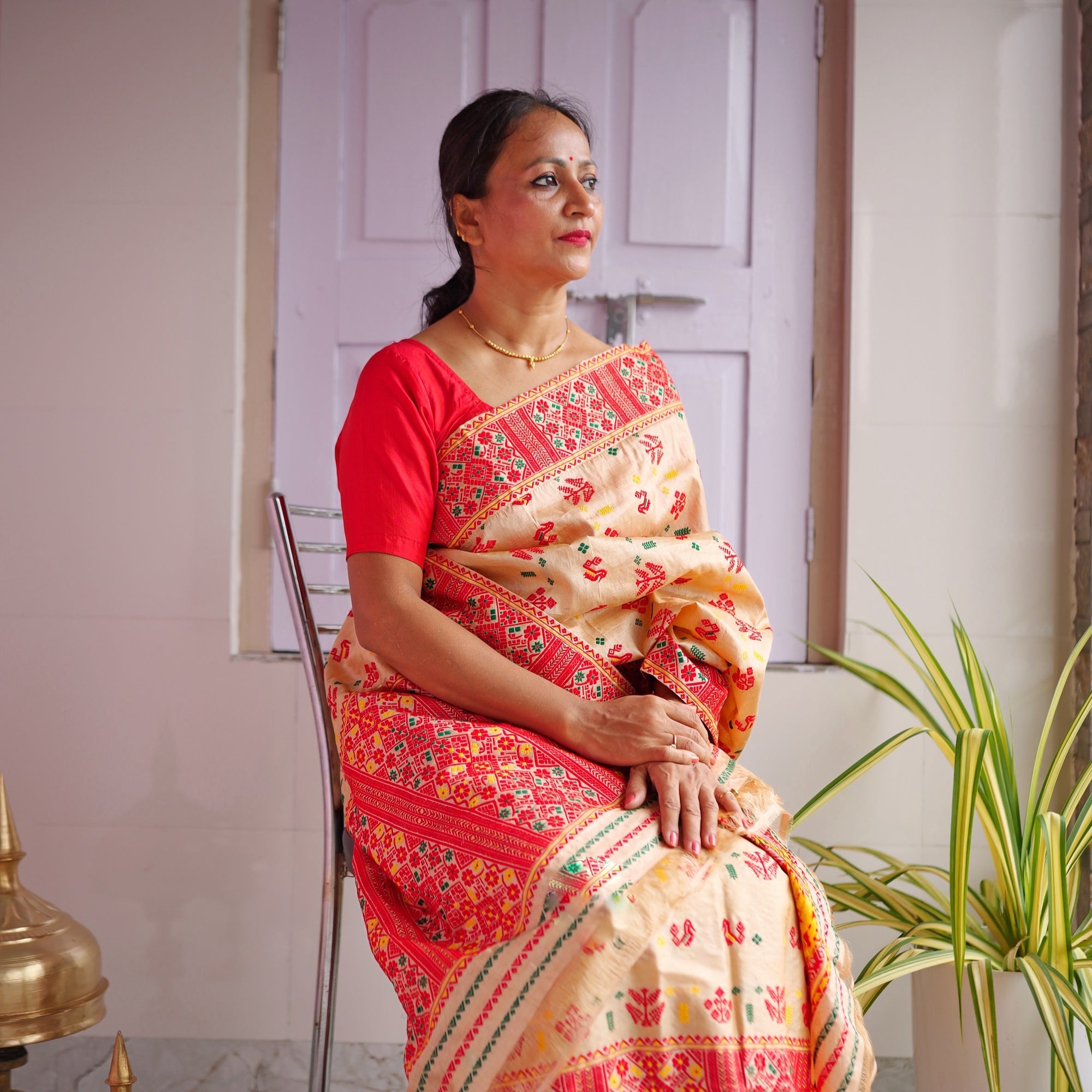 Seated on a chrome chair against a lilac panelled door, in a red blouse, looking to one side. A golden-beige drape with small red motifs, bordered and ended in dense red lattice, over a mekhela with a red lower panel.