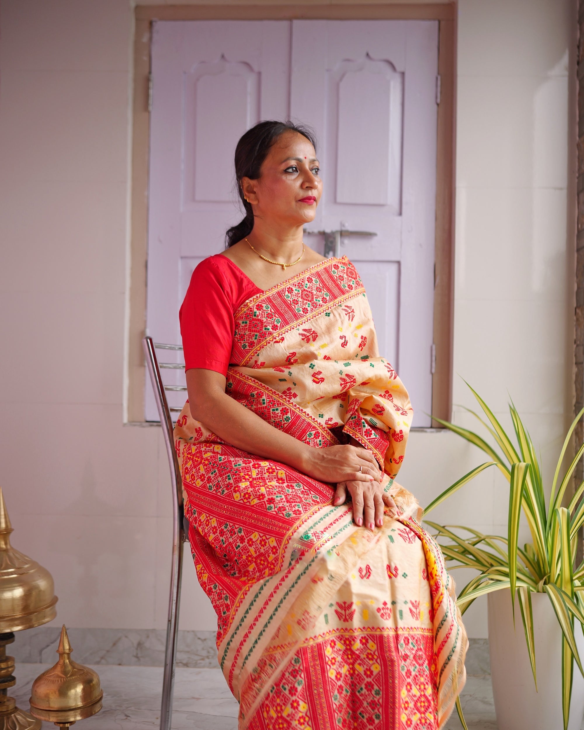 Seated on a chrome chair against a lilac panelled door, in a red blouse, looking to one side. A golden-beige drape with small red motifs, bordered and ended in dense red lattice, over a mekhela with a red lower panel.