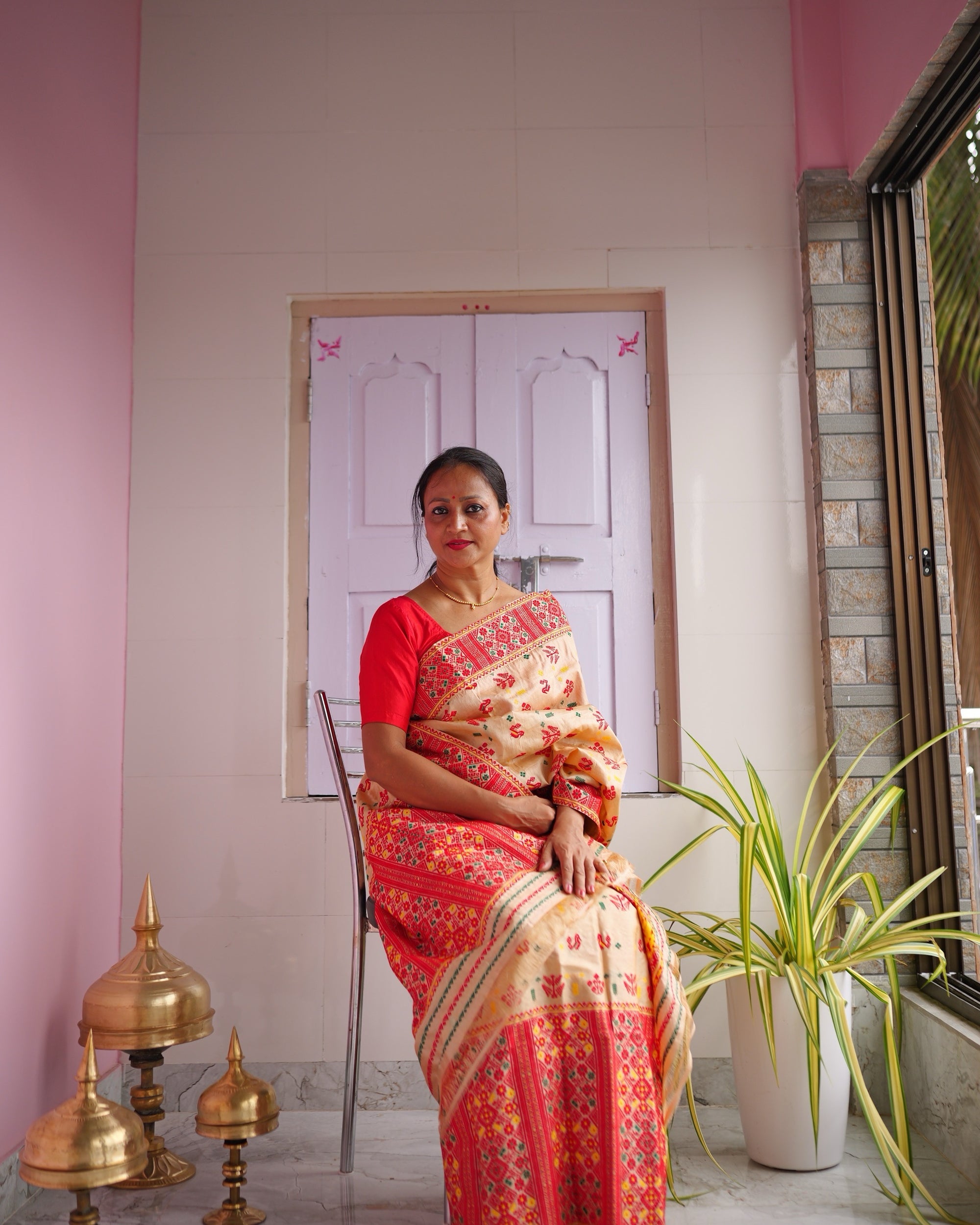 Seated on a verandah with pink walls and three brass xorai on the floor, the set seen full length.