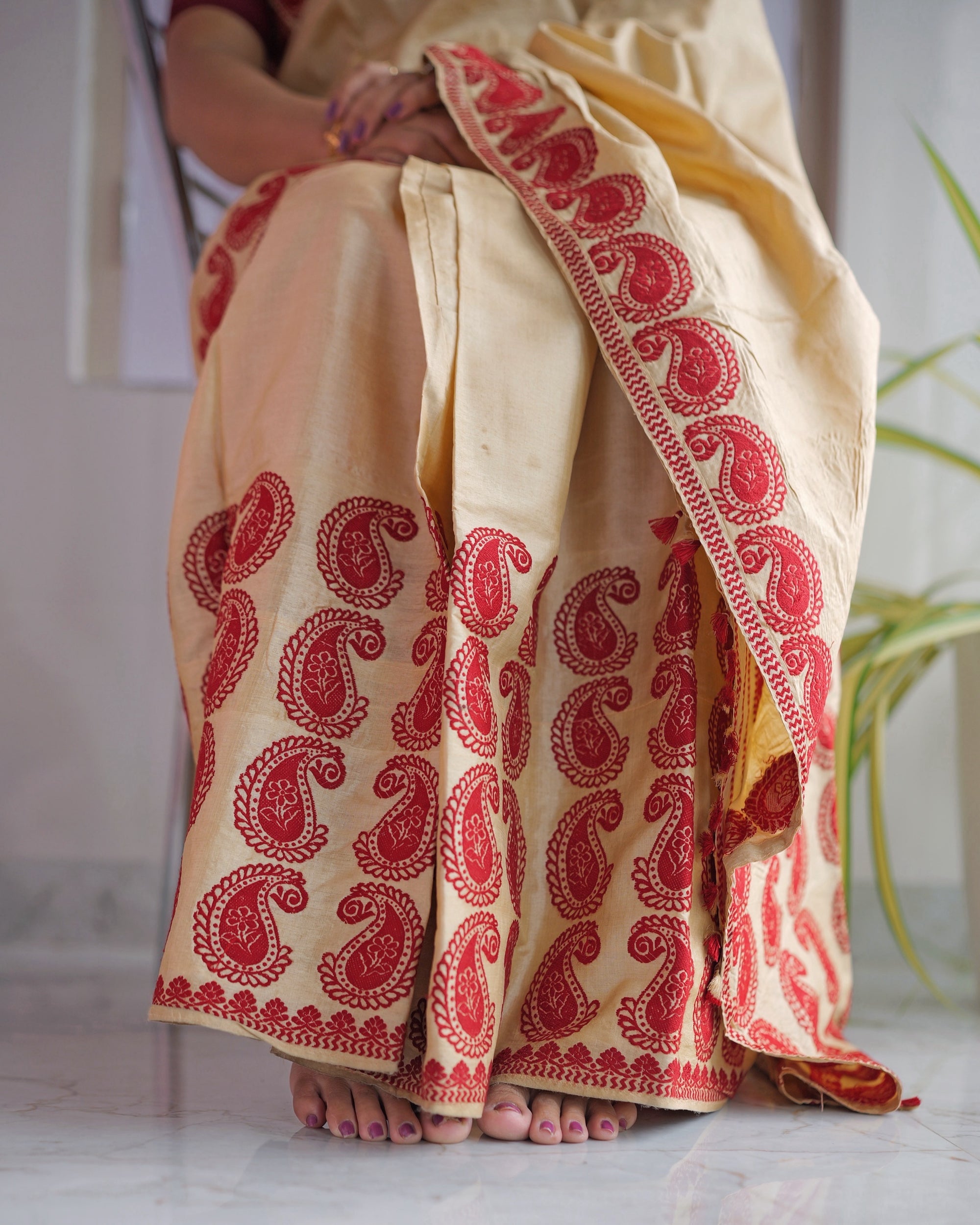 The hand-pleated mekhela falling to bare feet on a marble floor, red paisleys across the cream ground.