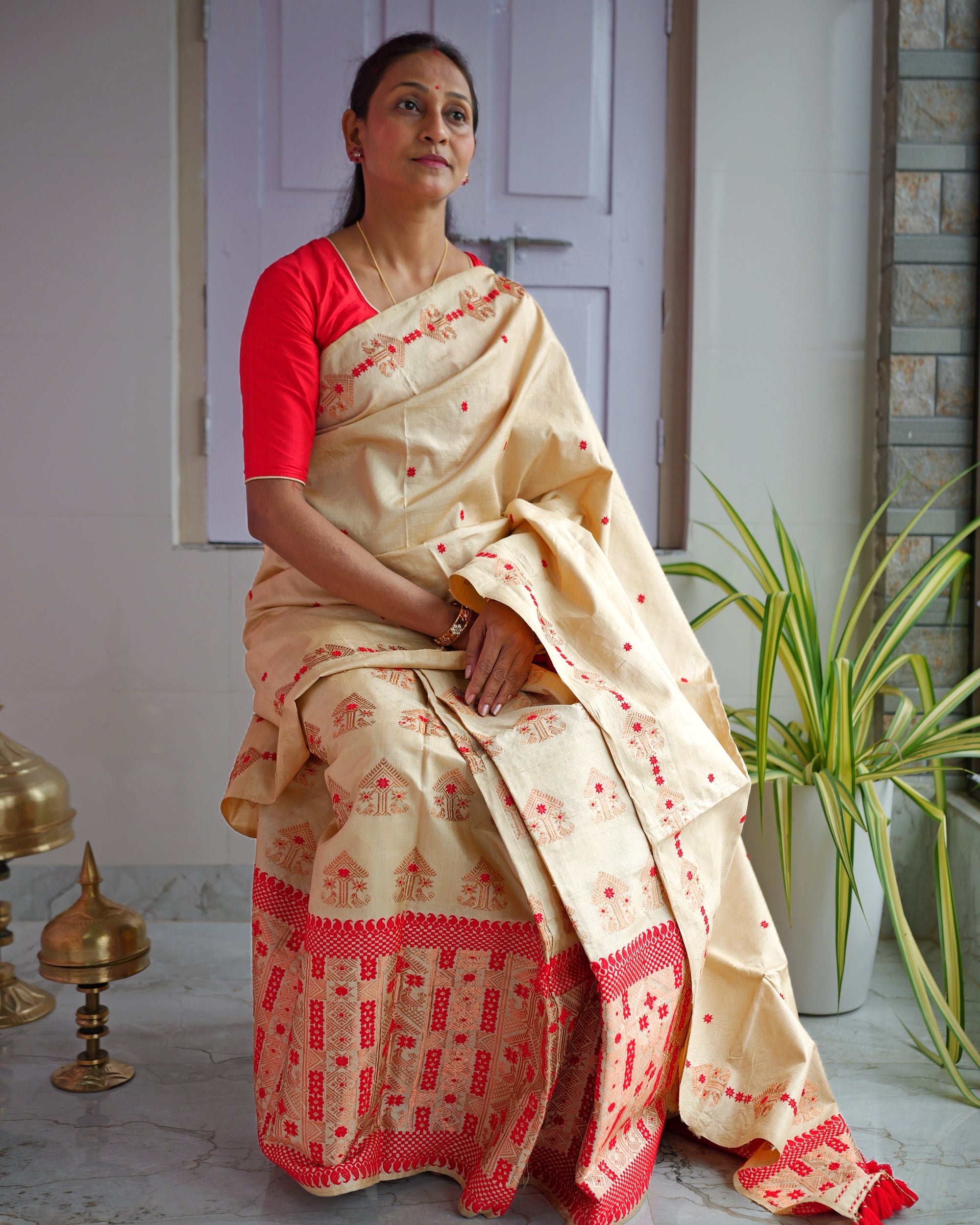Seated against a lilac panelled door in a red blouse, the whole set visible to the floor. A golden-beige drape dotted with small red florets and edged with copper-peach motifs, over a mekhela worked in red below the knee, red tassels at the end.