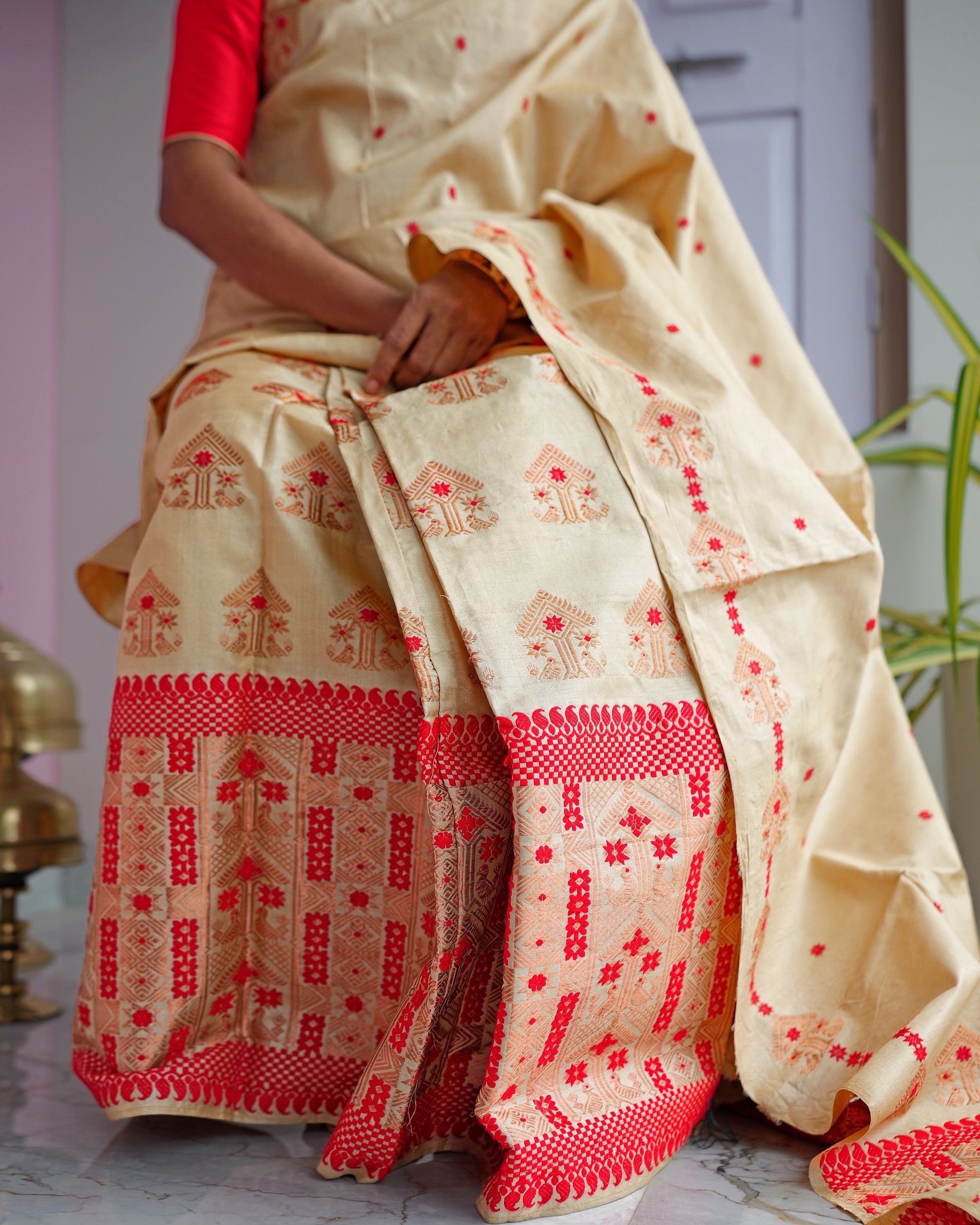 Seated, framed from the shoulder to the floor. Rows of copper-peach motifs on the mekhela above a red band of scallops and checks and a wide red panel; the drape falls to one side, its worked end on the floor.