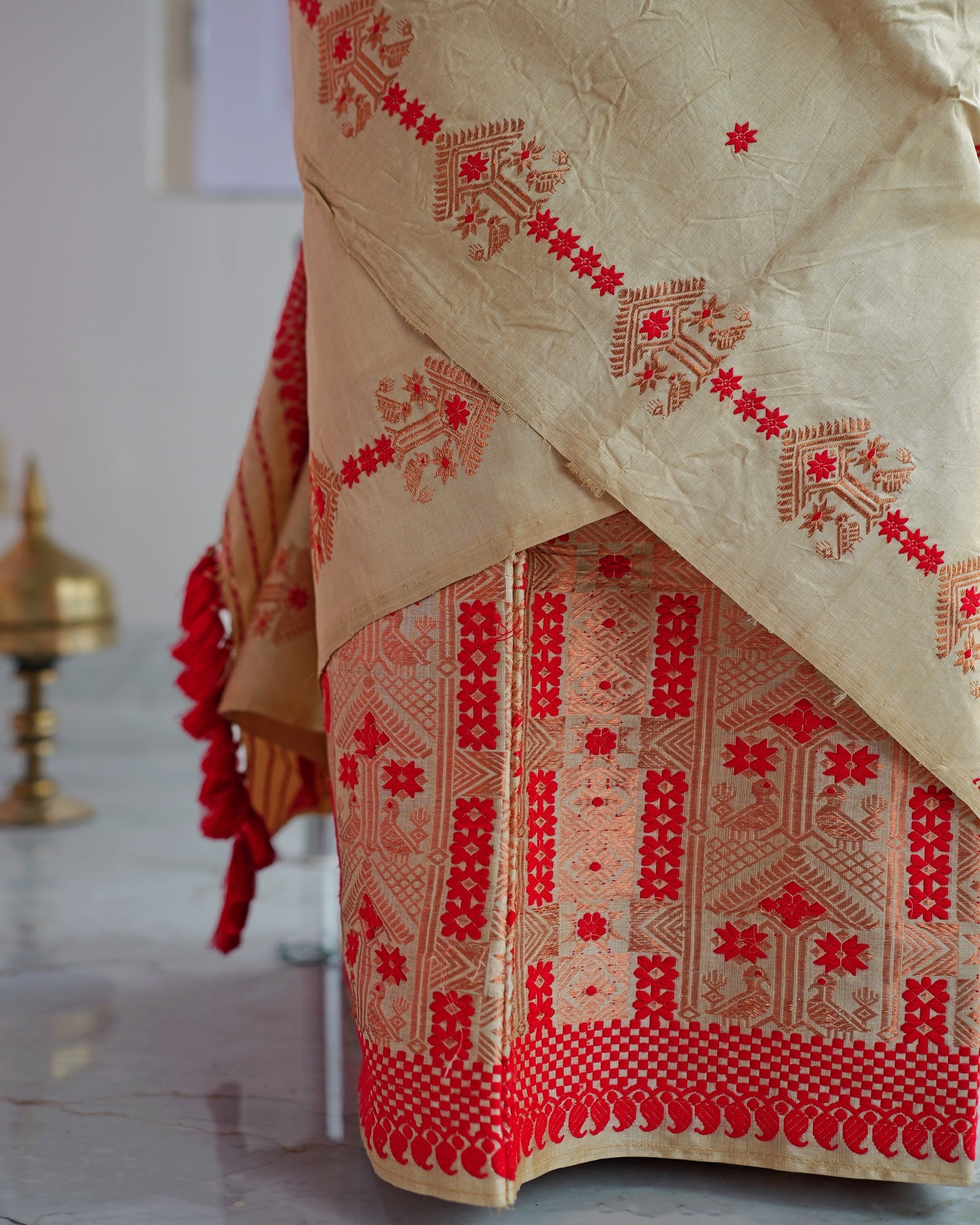 The drape hanging over the mekhela hem, close up. A line of copper-peach motifs linked by red florets crosses the beige drape; below, a red panel of columns, stars and paired copper-peach birds ends in red checks and scallops, with red tassels at the left.