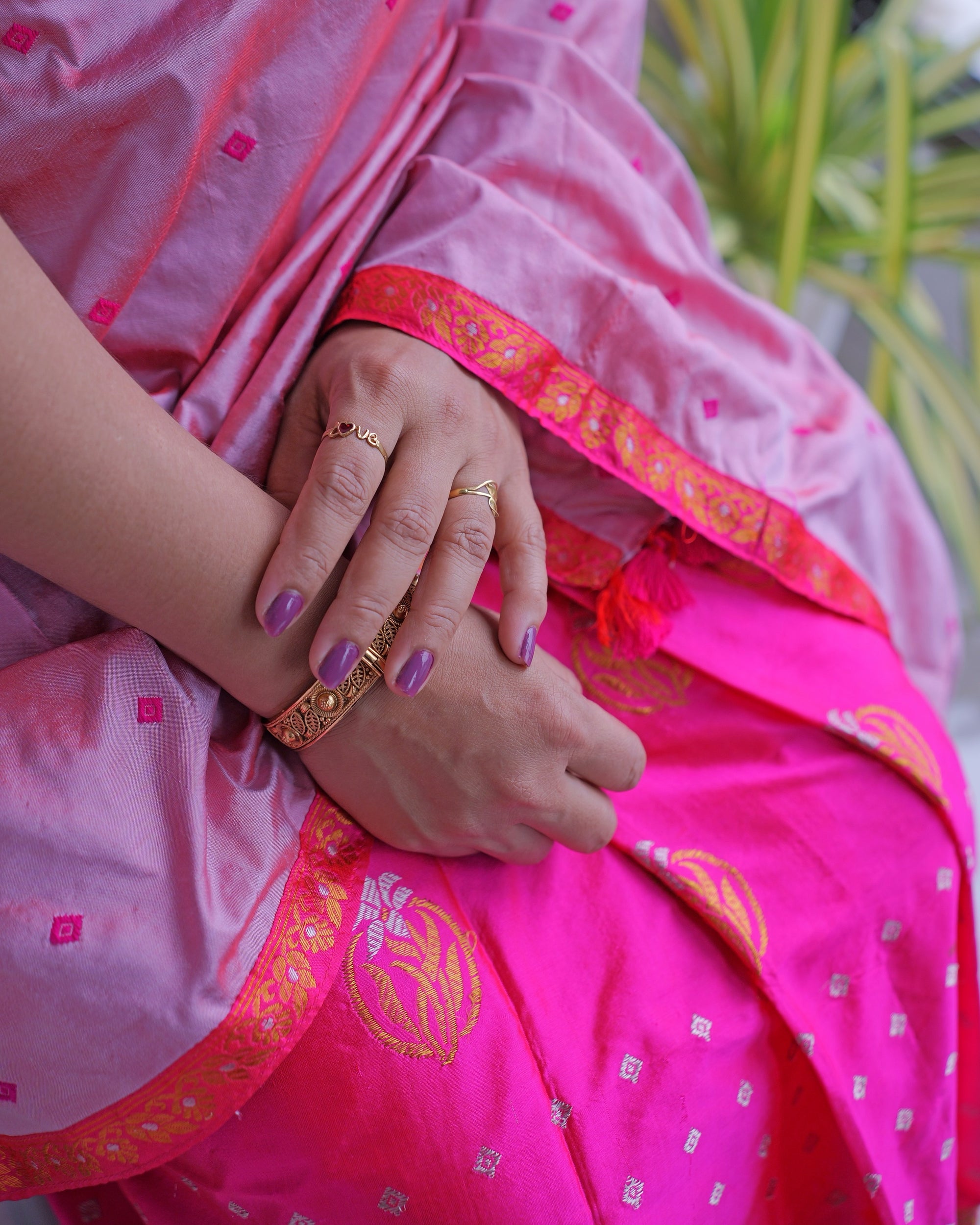 Hands resting in the lap, a gold bangle at the wrist, where the rose-pink drape meets the magenta mekhela along a golden-orange zari border.