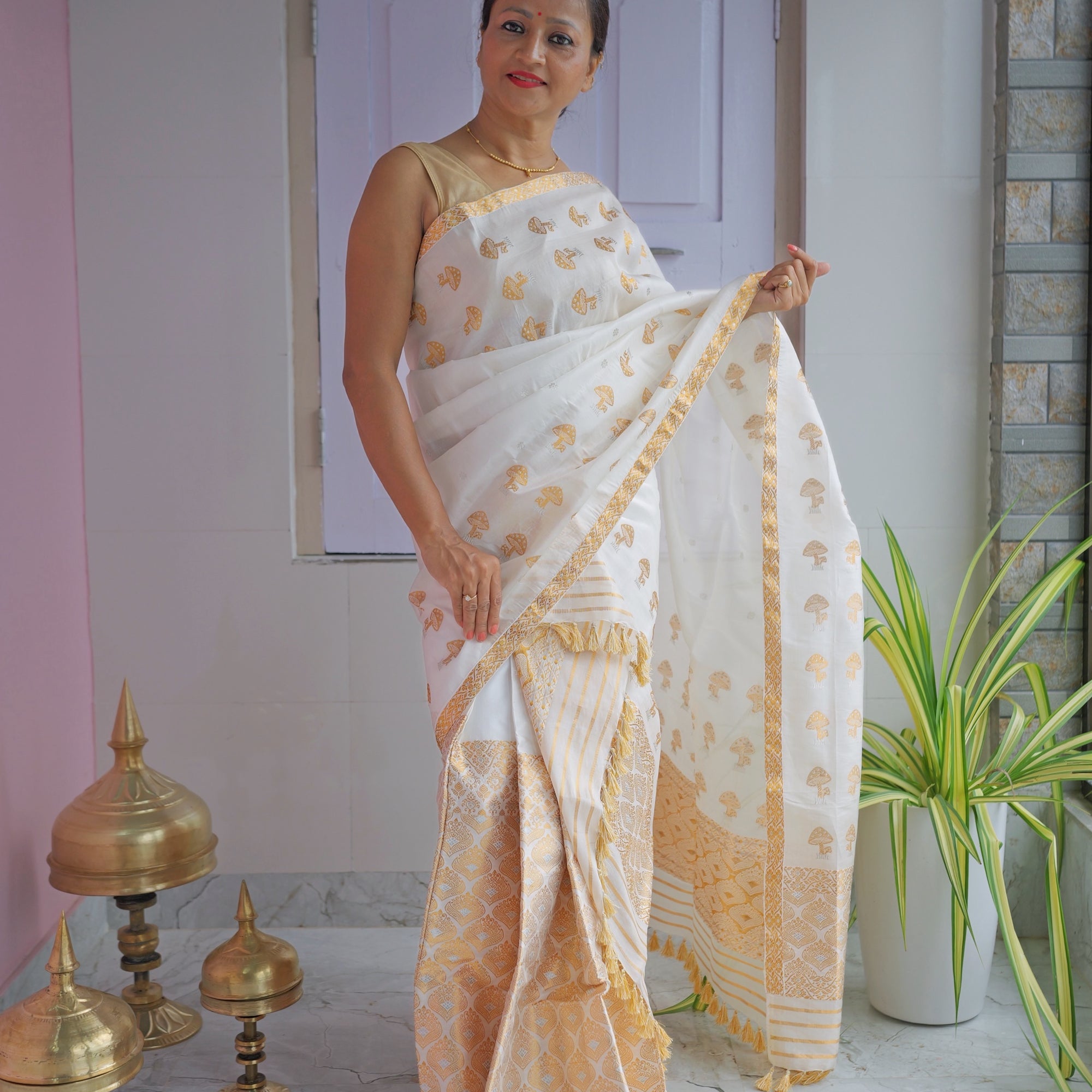 Standing against a panelled door, the whole set visible. An ivory body scattered with small gold mushroom motifs, over a mekhela hem worked in dense gold zari.
