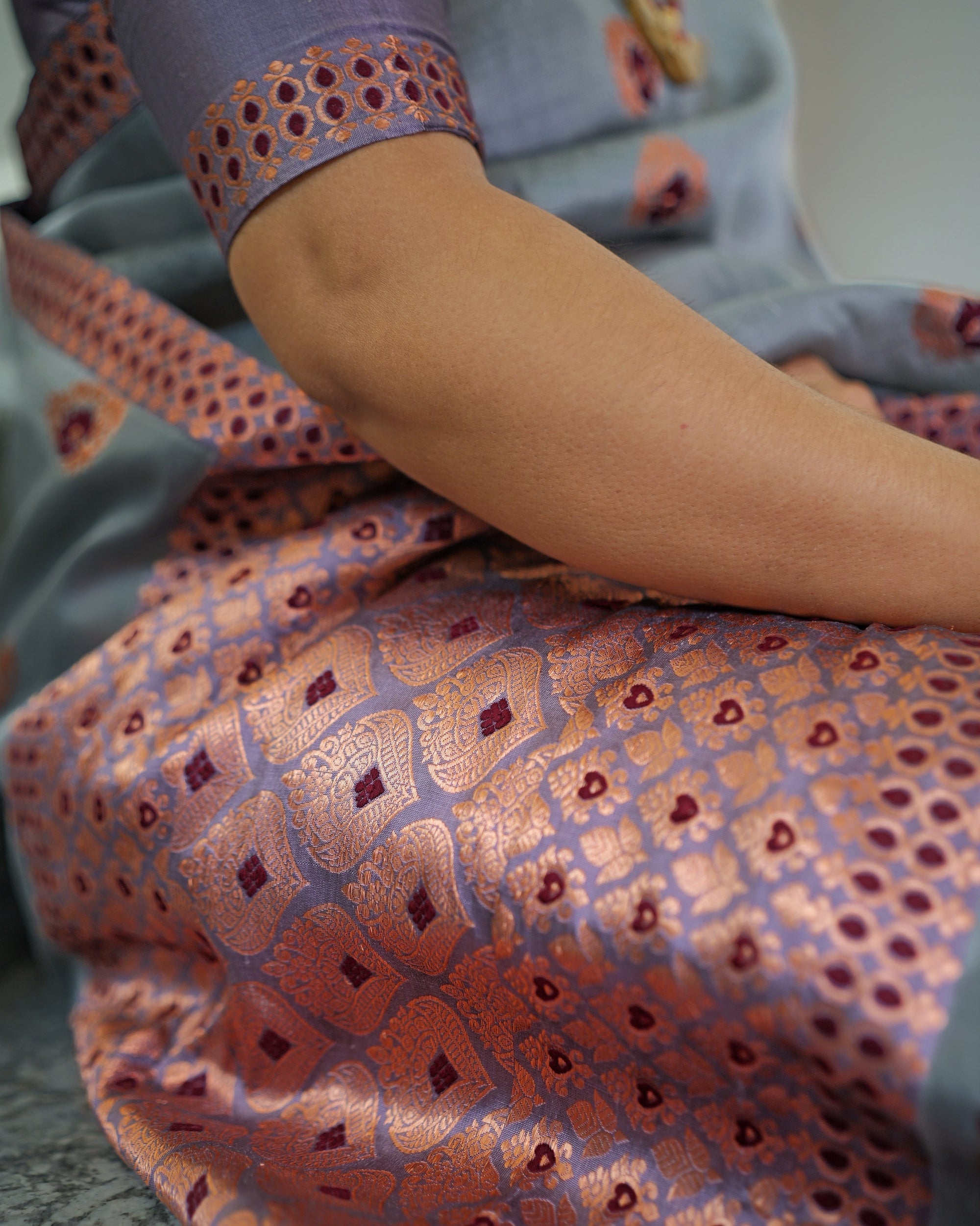 The mekhela close up: arched cells of copper zari on lilac, each centred on a small maroon diamond, beside a finer pattern of copper florets with maroon hearts, under a forearm and a lilac sleeve edged in copper.