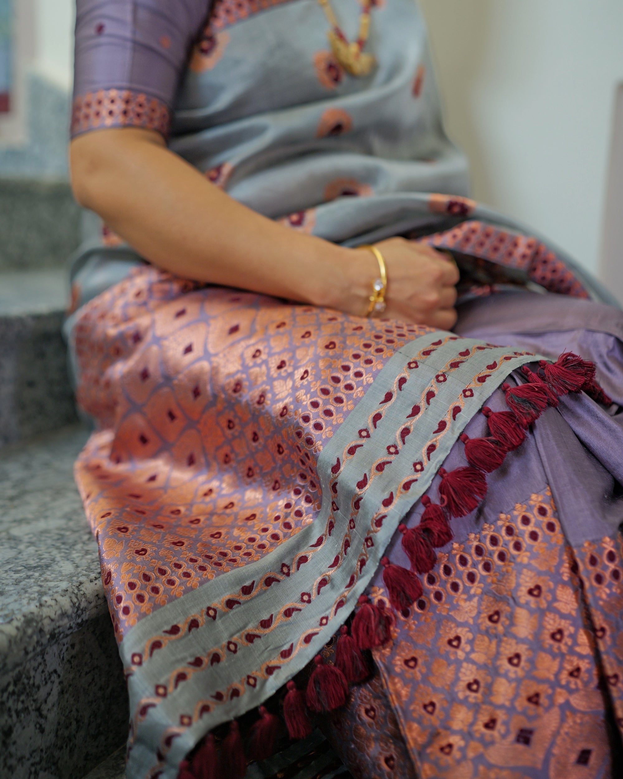 The sador end across the lap, grey bands lined with small maroon marks between copper stripes, maroon tassels falling over the copper lattice of the mekhela, a gold bangle at the wrist.