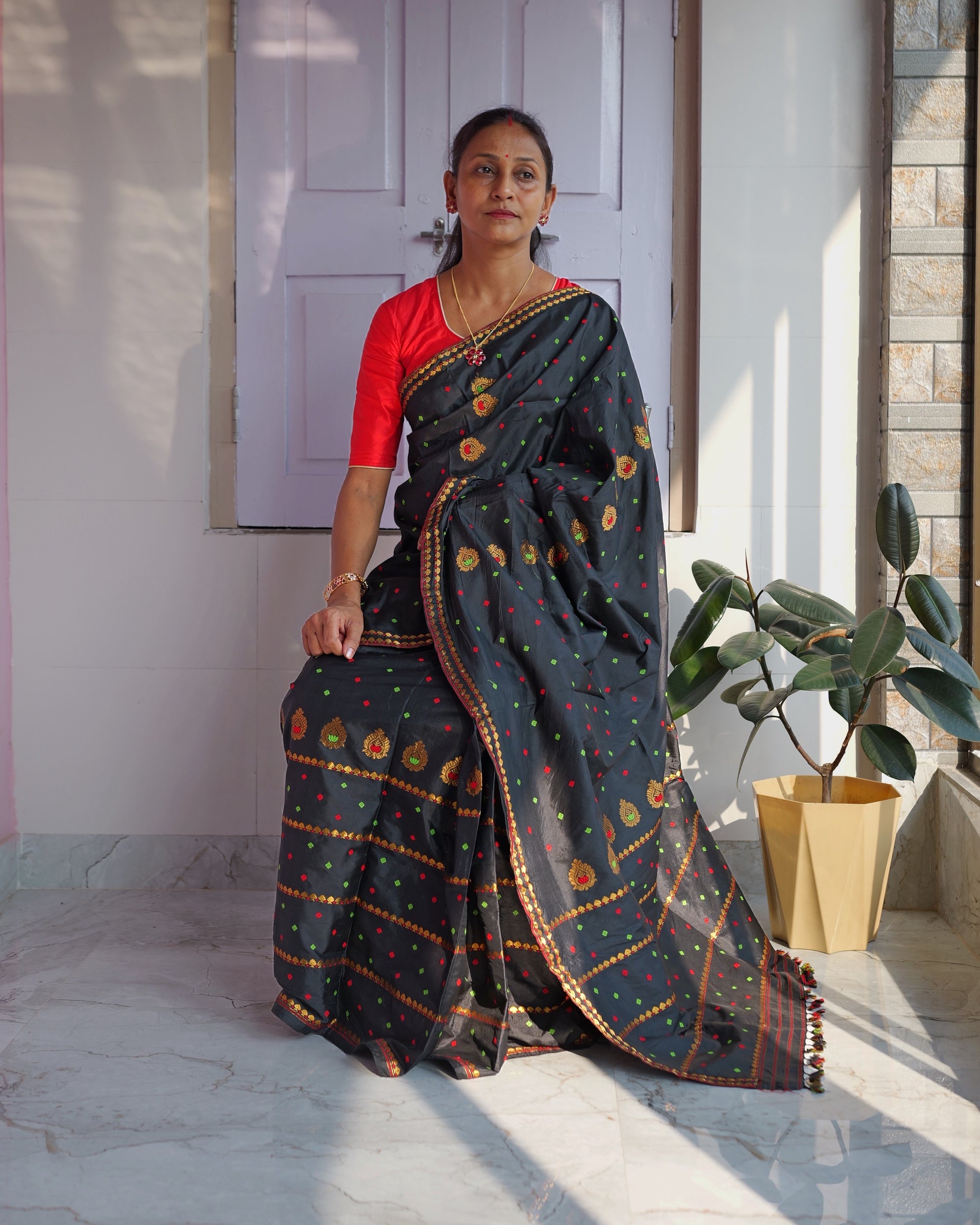Seated against a pale panelled door, sunlight falling across the floor, the whole set visible. A black drape over a red blouse, scattered with red and green squares and round gold flowers, the mekhela crossed by gold bands, small coloured tassels at the sador end.