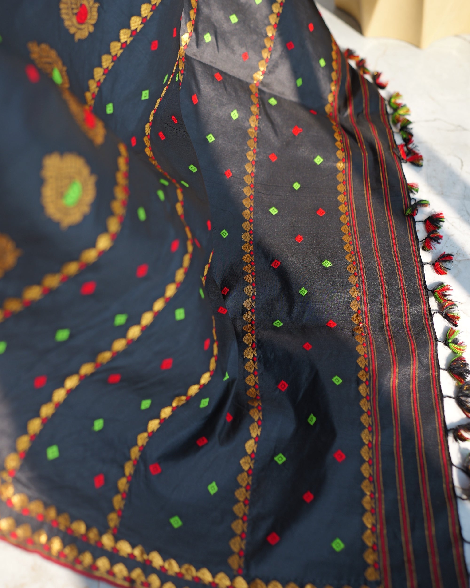 The sador end in raking sun: red and green squares, round gold flowers and gold fan borders, then red and gold stripes and small black, red and green tassels flecked with gold along the edge.