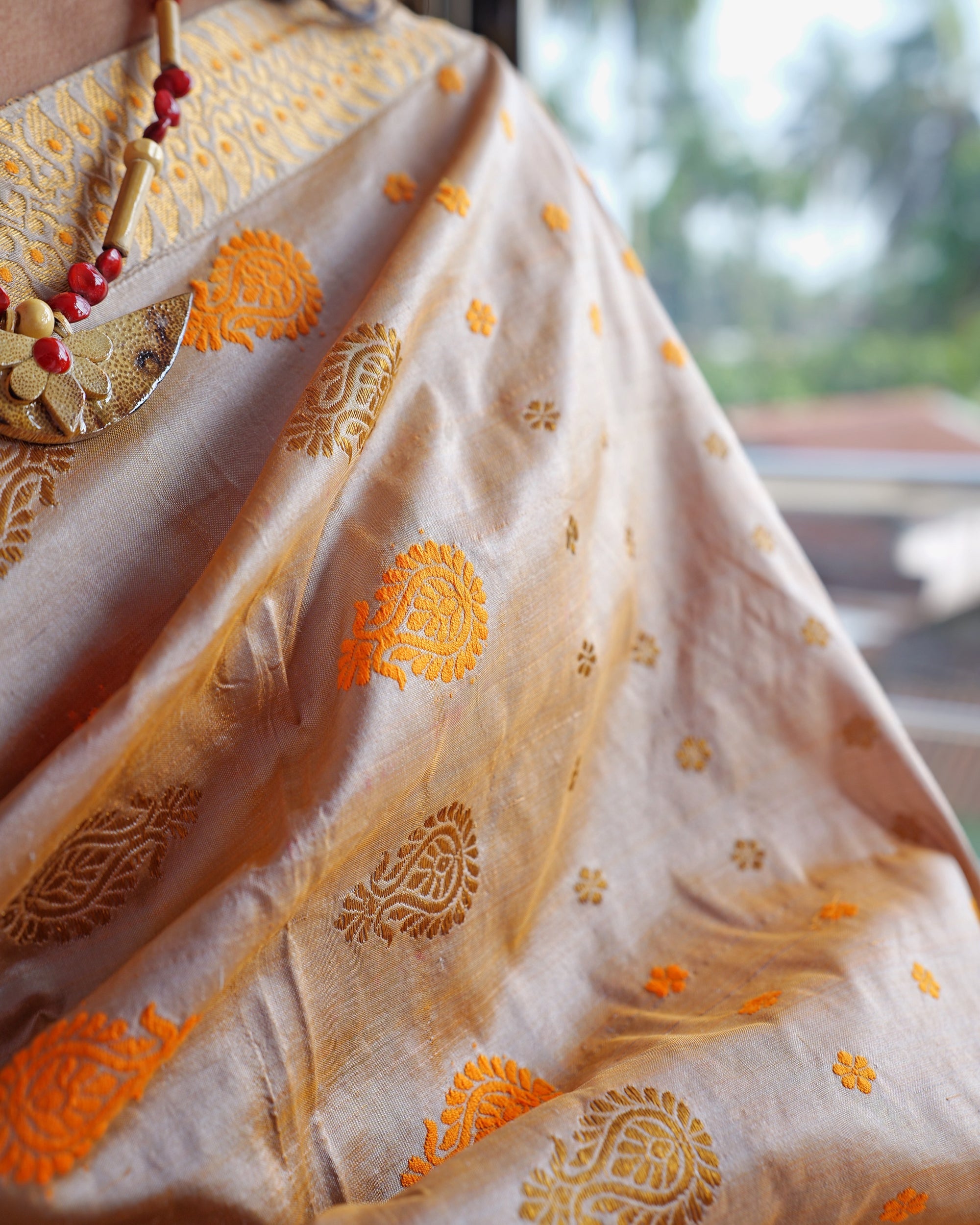 The drape over the shoulder close up: the same leafy paisley woven in saffron and in gold-toned zari, small florets between, under a broad zari border and a bead necklace with a crescent pendant.
