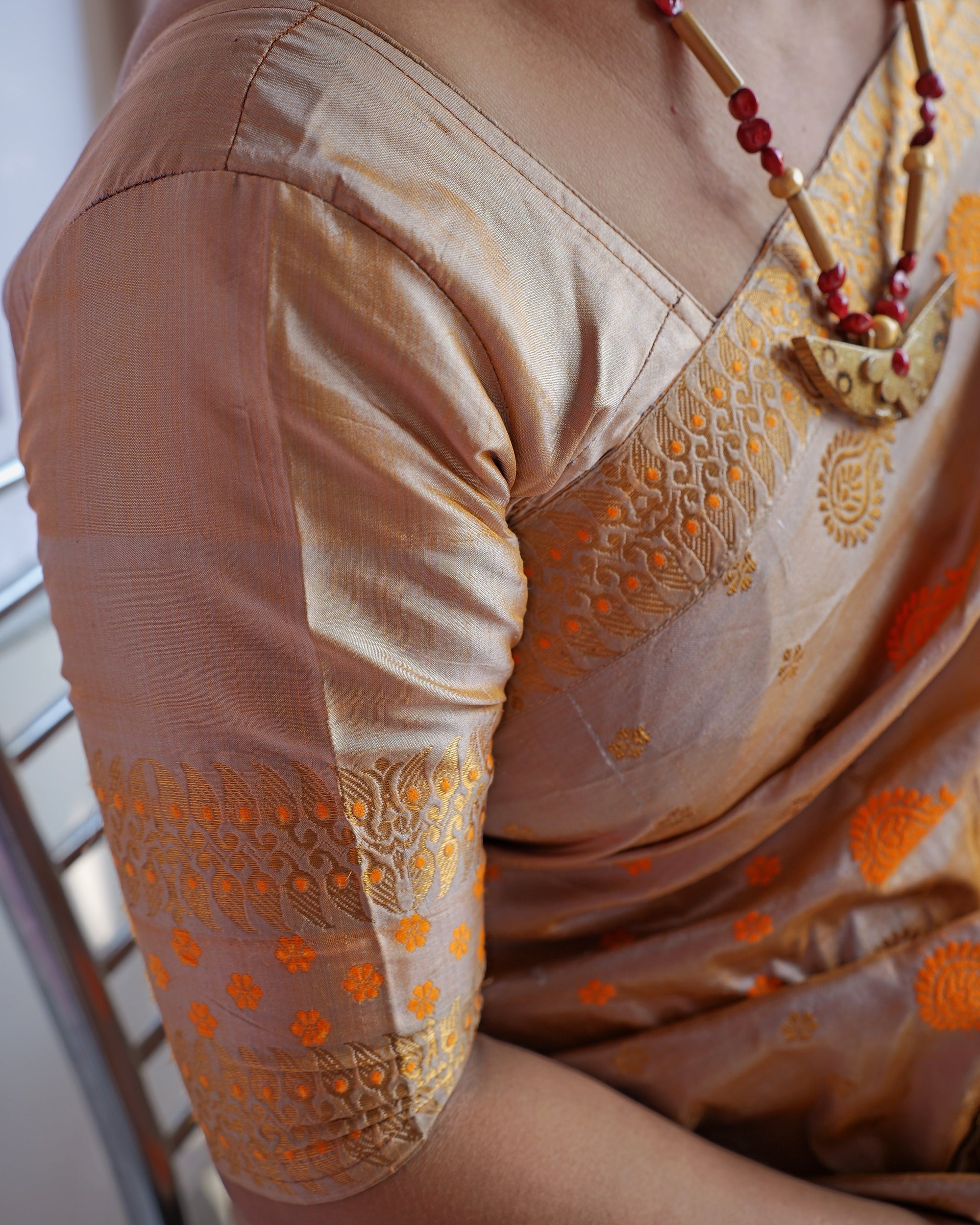 The shoulder and sleeve: a beige blouse whose sleeve is edged with zari scrolls and saffron florets, the sador's zari border across the chest, a red bead necklace with a crescent pendant.