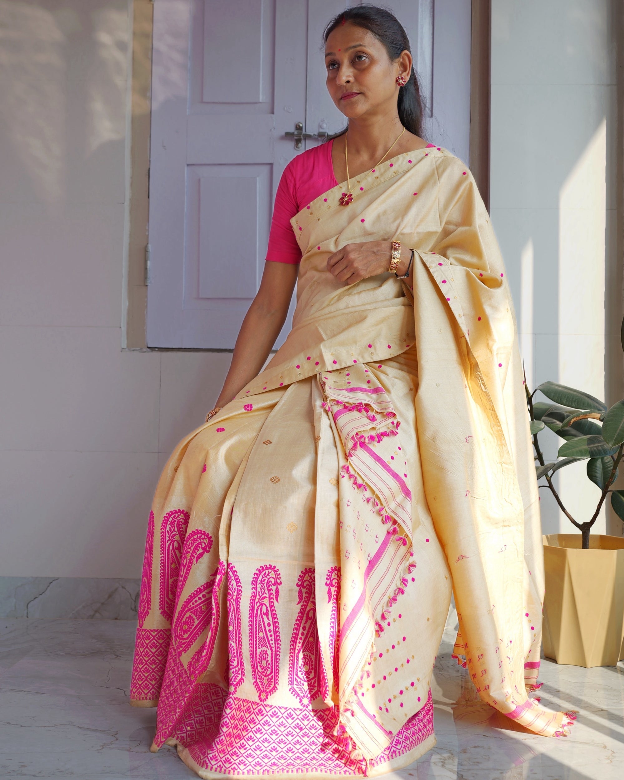 Seated against a lilac panelled door beside a potted plant, the whole set visible. A golden-cream drape dotted with pink and gold, over a mekhela hem of tall rani pink paisleys and a deep pink border, worn with a rani pink blouse.