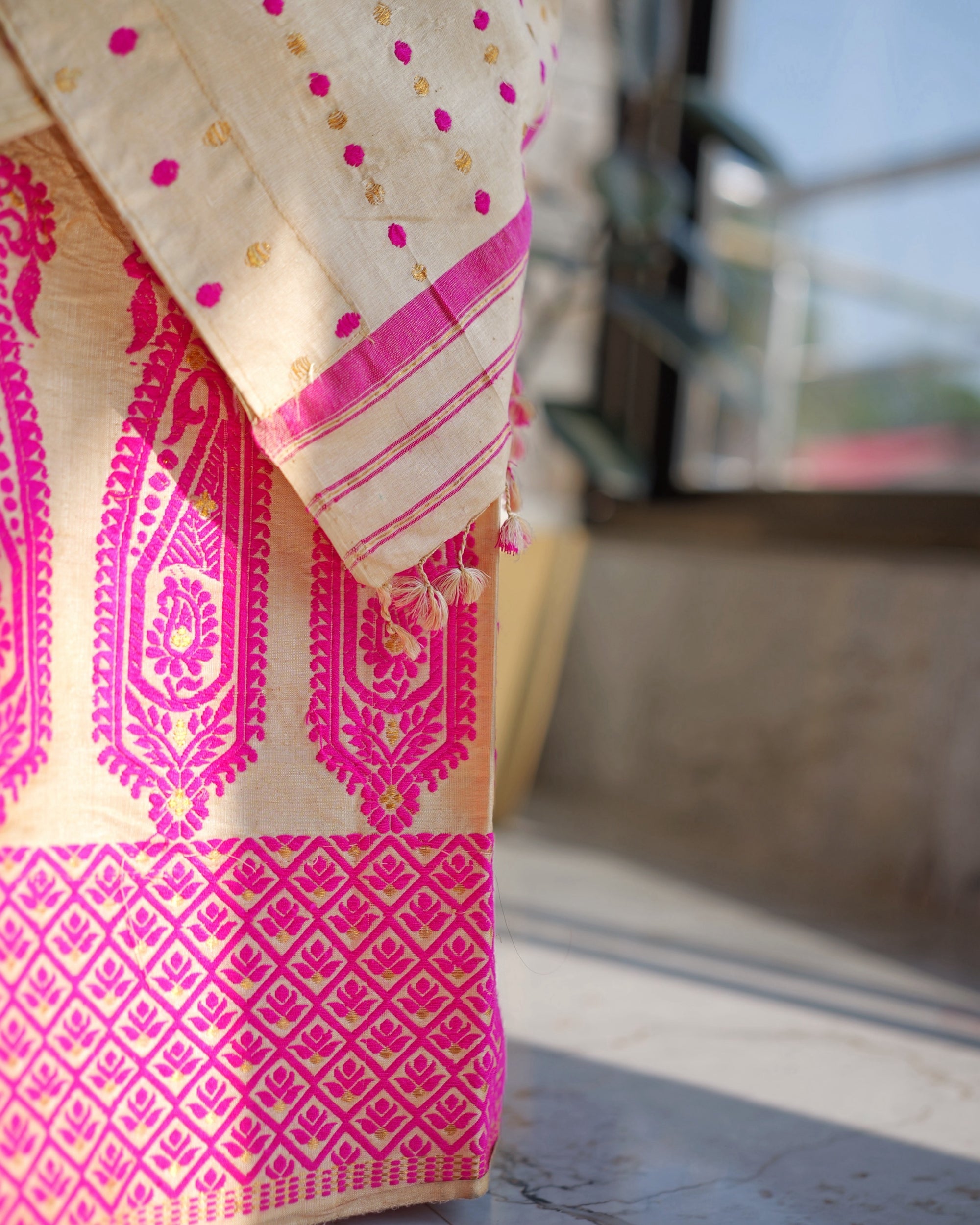 The lower mekhela in window light. Tall pink paisleys above a border of pink diamonds, with the striped end of the sador and its small cream and pink tassels hanging over them.