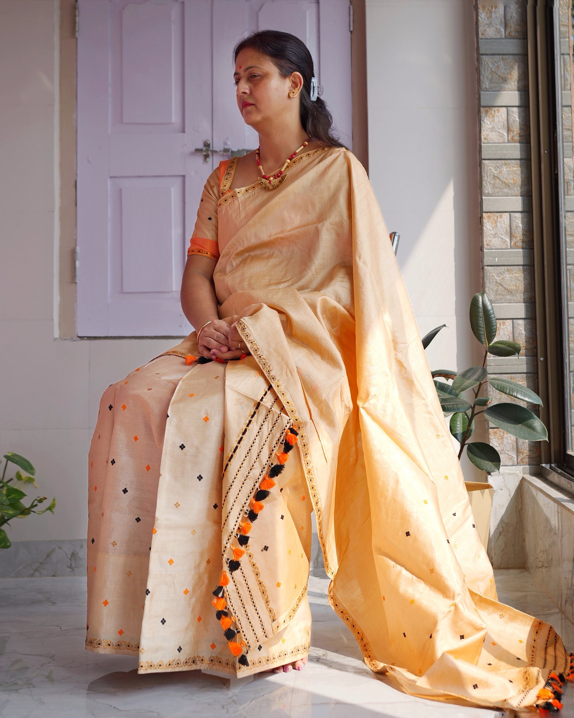 Seated on a chair against a lilac panelled door, looking away to the left, the whole set visible. A golden-cream body scattered with small orange and black motifs, and the checked end of the sador hanging from the lap to the floor in a row of orange and black tassels.