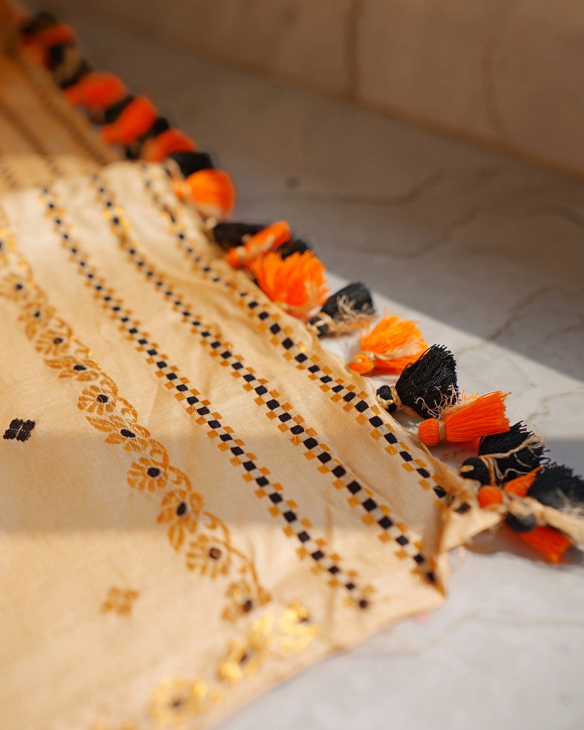The end of the sador laid on a sunlit marble floor. Lines of tiny black and gold checks and a vine of small gold flowers, finished with a row of orange and black tassels on twisted cords.