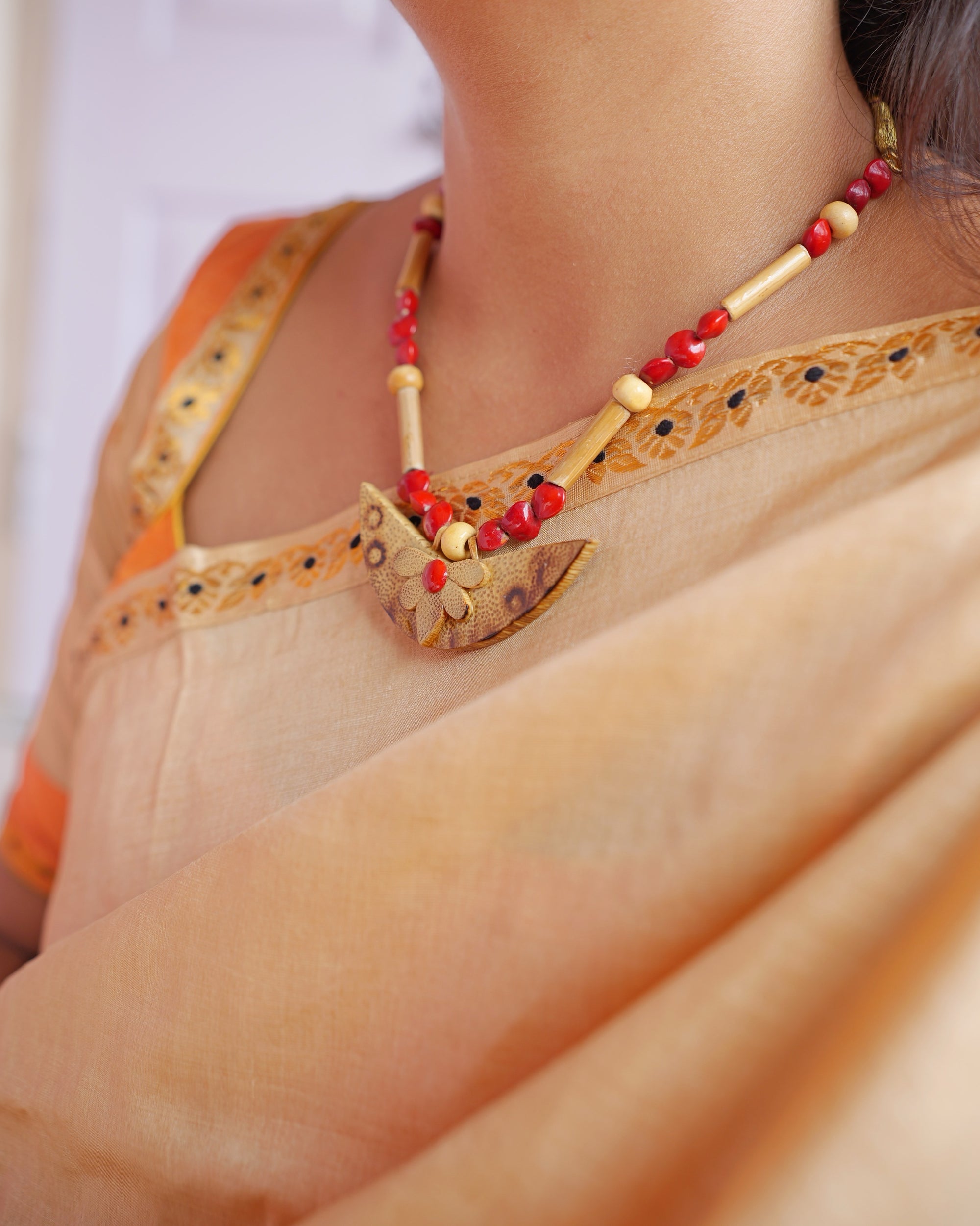 The neckline close up: a golden blouse with an orange sleeve, the sador's edge of small gold flowers with black centres crossing below the collarbone, and a necklace of red and pale beads on a crescent pendant.