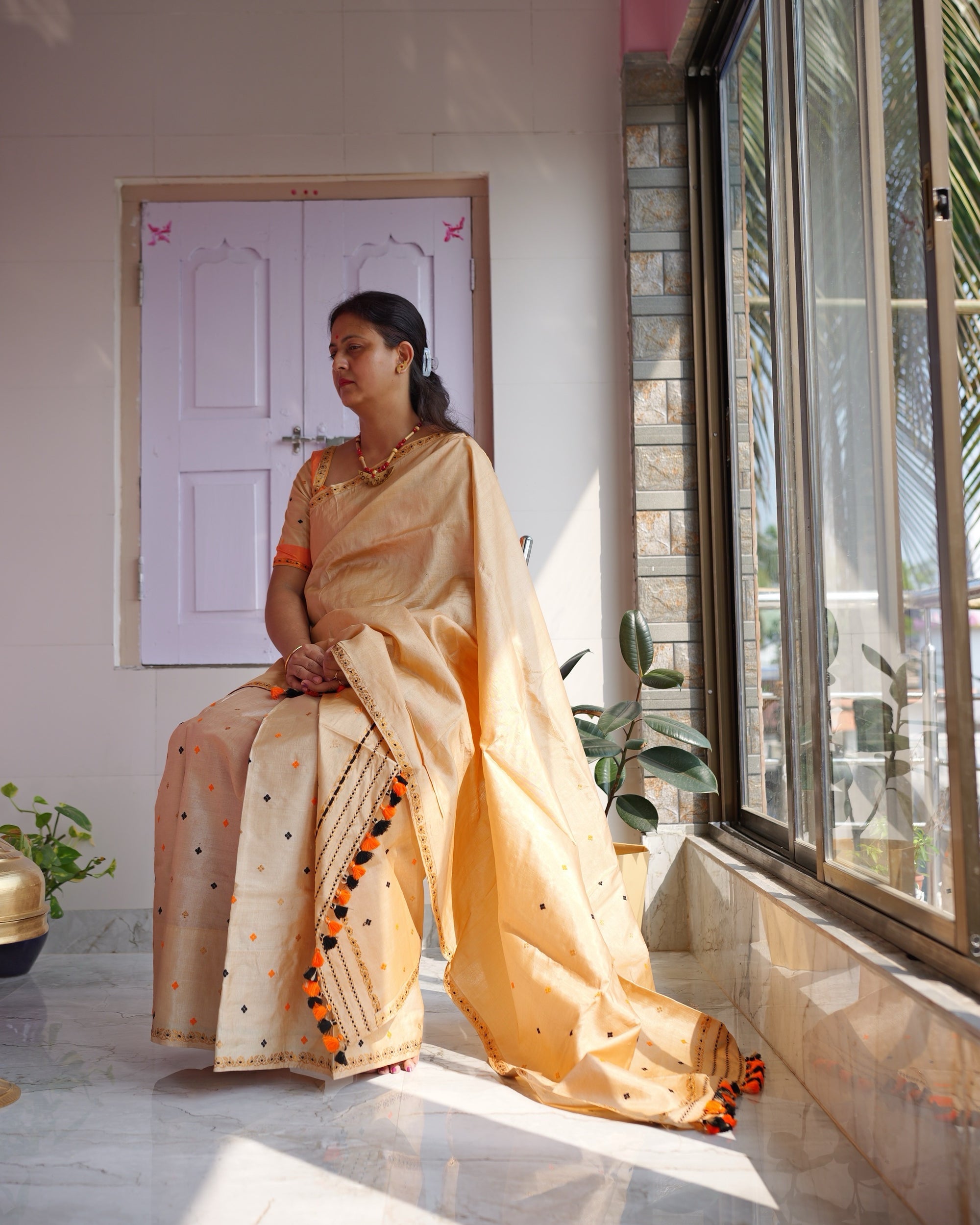 Seated by a tall window on a verandah, a pink wall above and a lilac door behind, the set seen full length with the tasselled end of the sador trailing on the floor.