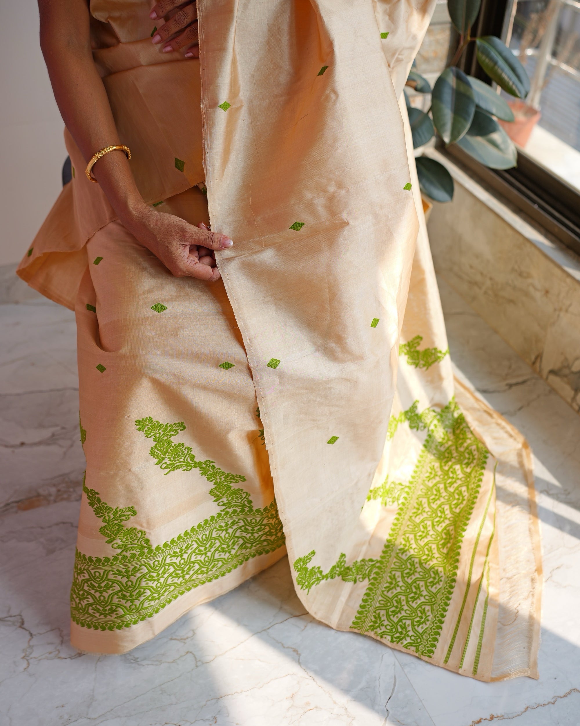 Seated, framed from the waist down in strong sunlight, a hand holding the edge of the sador. Green scrollwork rises in steps from the hem of the mekhela, and the sador falls beside it to a marble floor, its end worked in green vines and stripes.