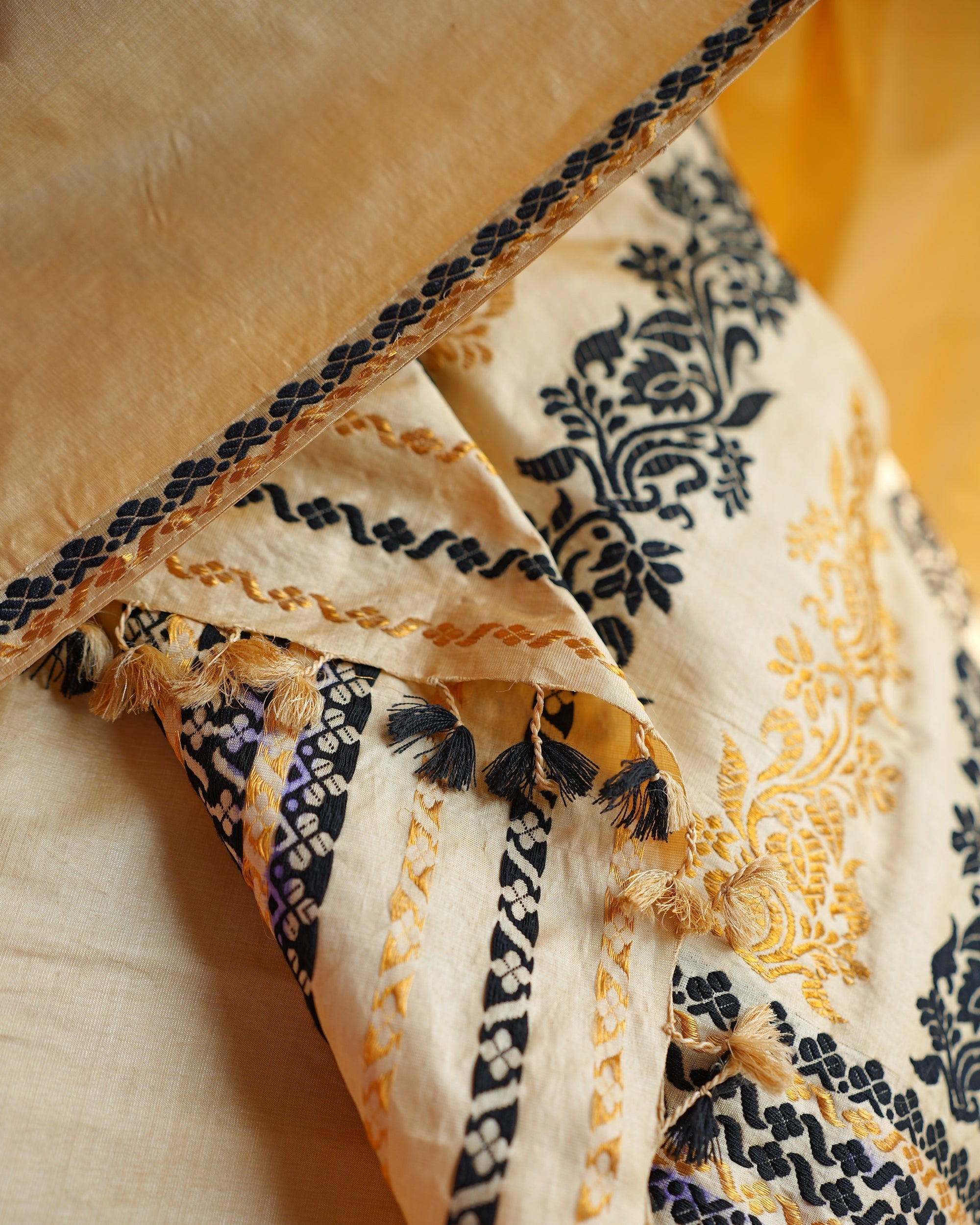 The sador end folded over, close up. Black and gold flowering creepers, black bands patterned with small cream florets, wavy gold lines, and tassels in black and in gold.
