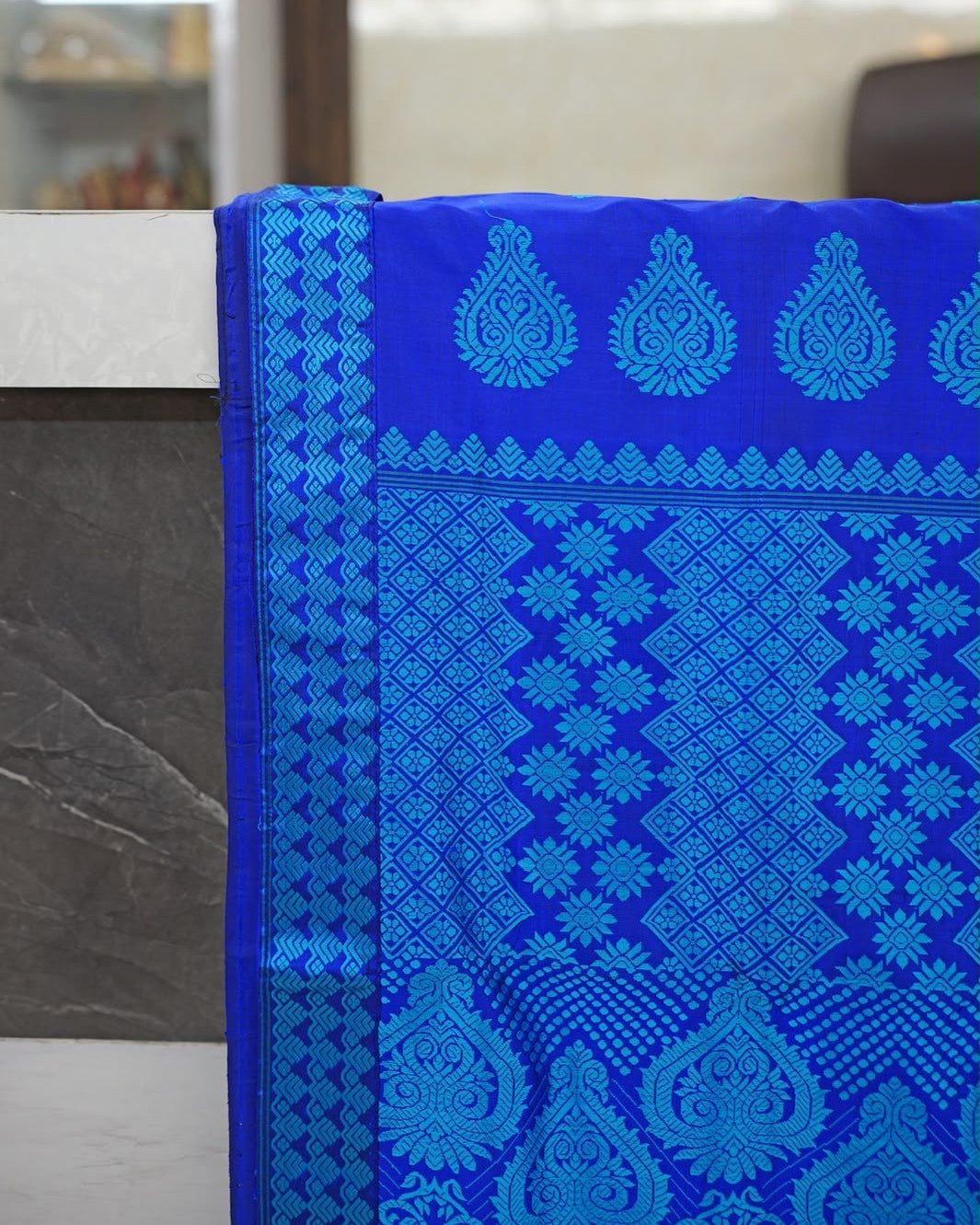 The pallu hung over the edge of a stone counter and seen straight on: a turquoise chevron border down the left side, feathered teardrop medallions across the top, then columns of stepped diamonds and star-shaped flowers above large heart-shaped medallions.