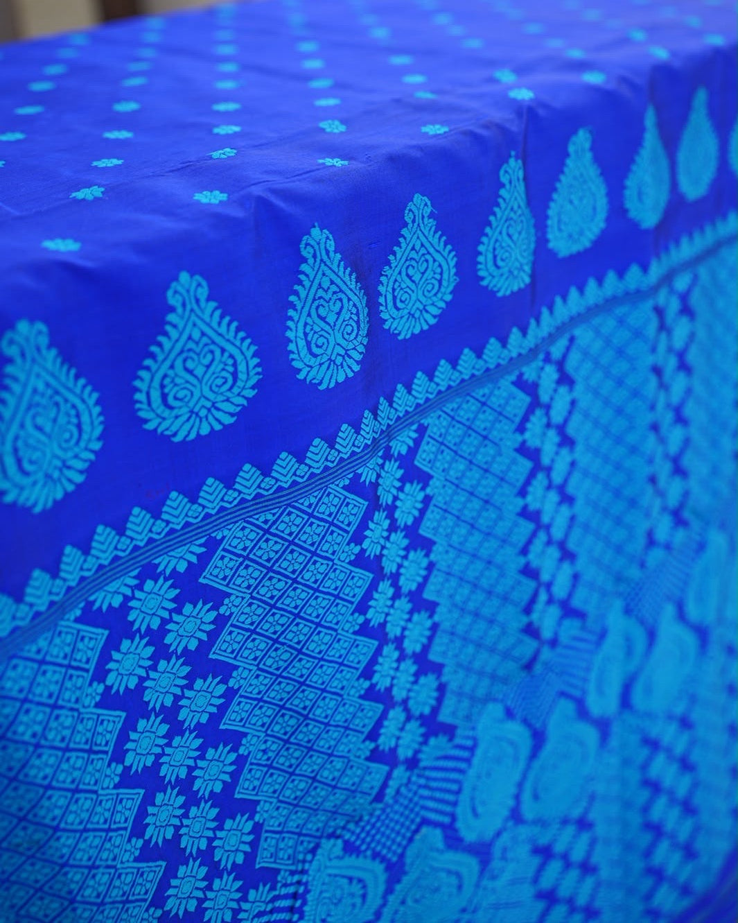 The saree laid along a table and seen at an angle: small turquoise flowers on the royal-blue body give way to a row of feathered teardrop medallions, a band of stepped triangles and the diamond panels of the pallu.