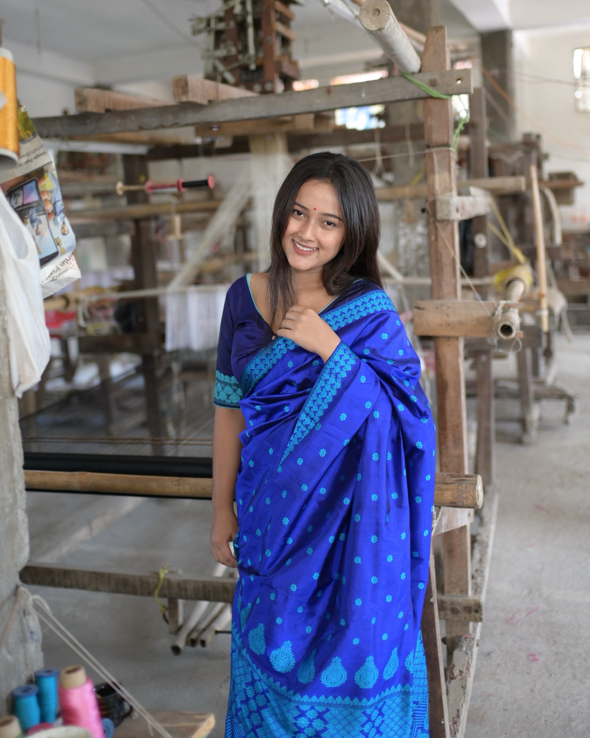 Smiling beside a handloom, one hand at the shoulder: the pallu over the shoulder, turquoise flowers on royal blue and the turquoise border along its edge.