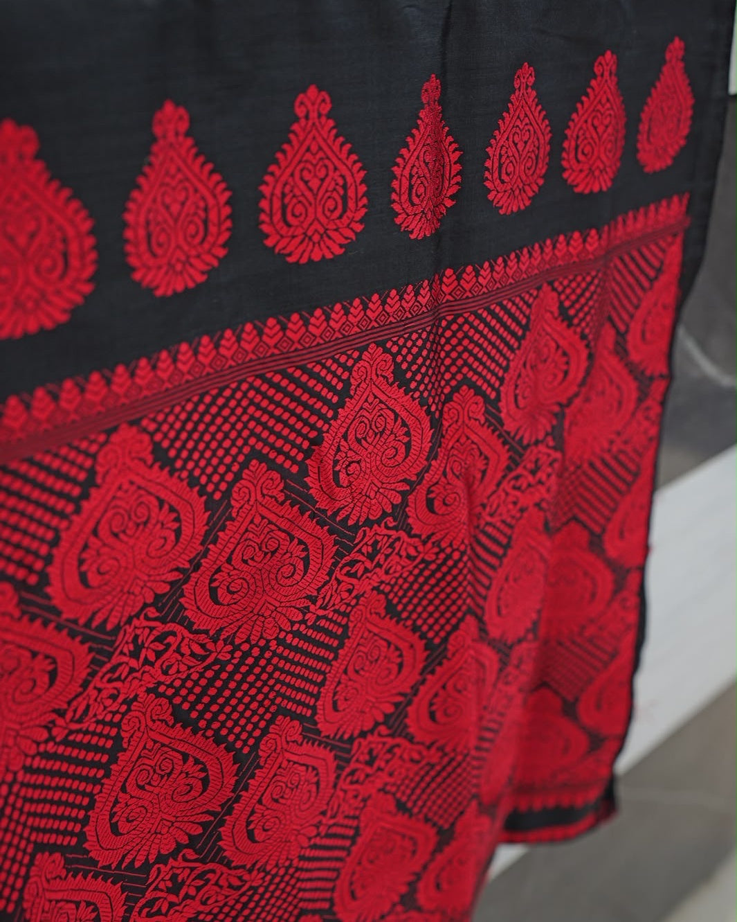 The pallu draped over the corner of a counter and seen at an angle, one side falling free: a row of red teardrop medallions on black, a narrow band of small leaves, then a dense field of red scrollwork medallions and dotted chevrons, softening out of focus to the right.