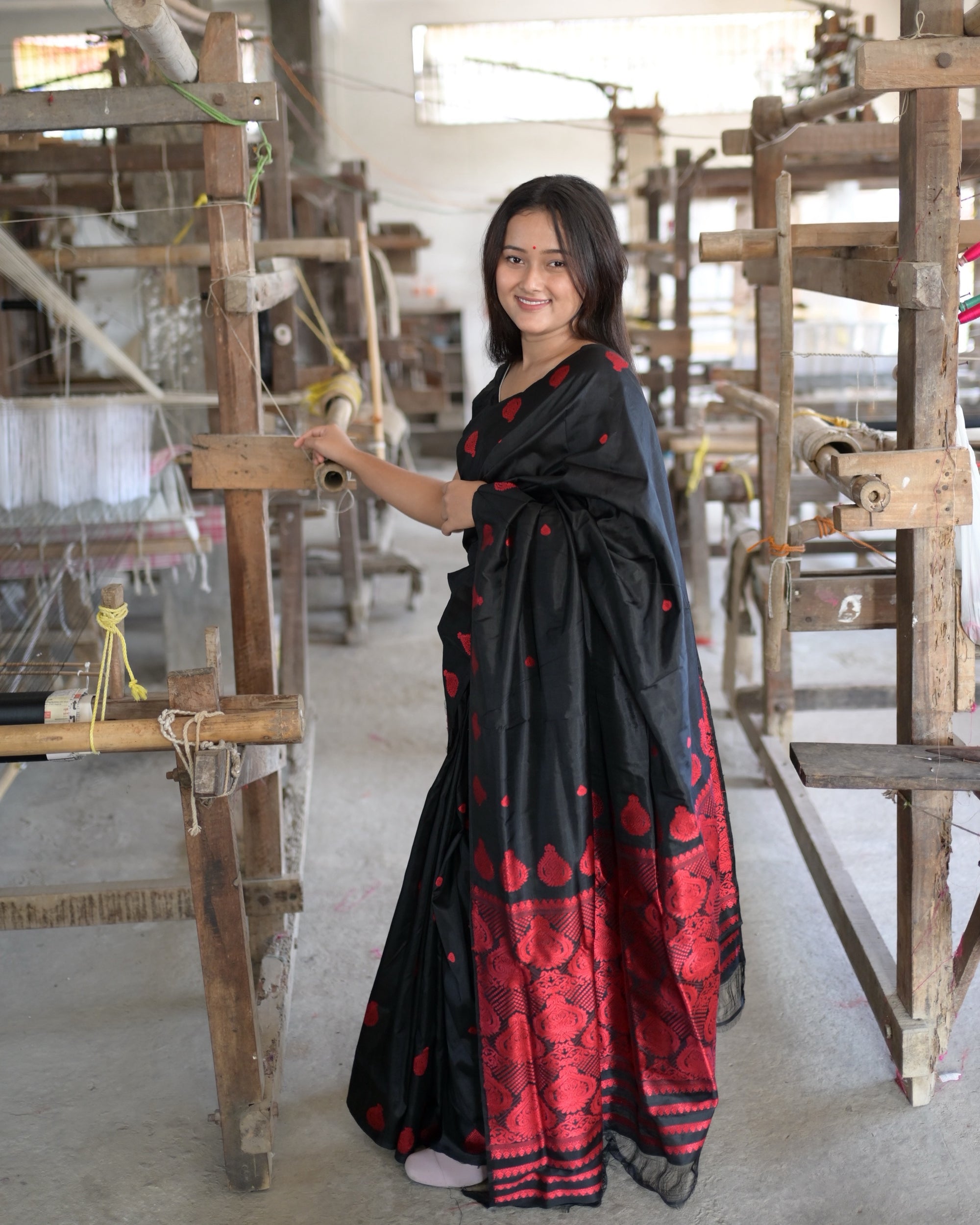 Standing at a wooden loom in a weaving hall, one hand on its beam: small red drops over the black body and the pleats falling to a red field of medallions above the hem.