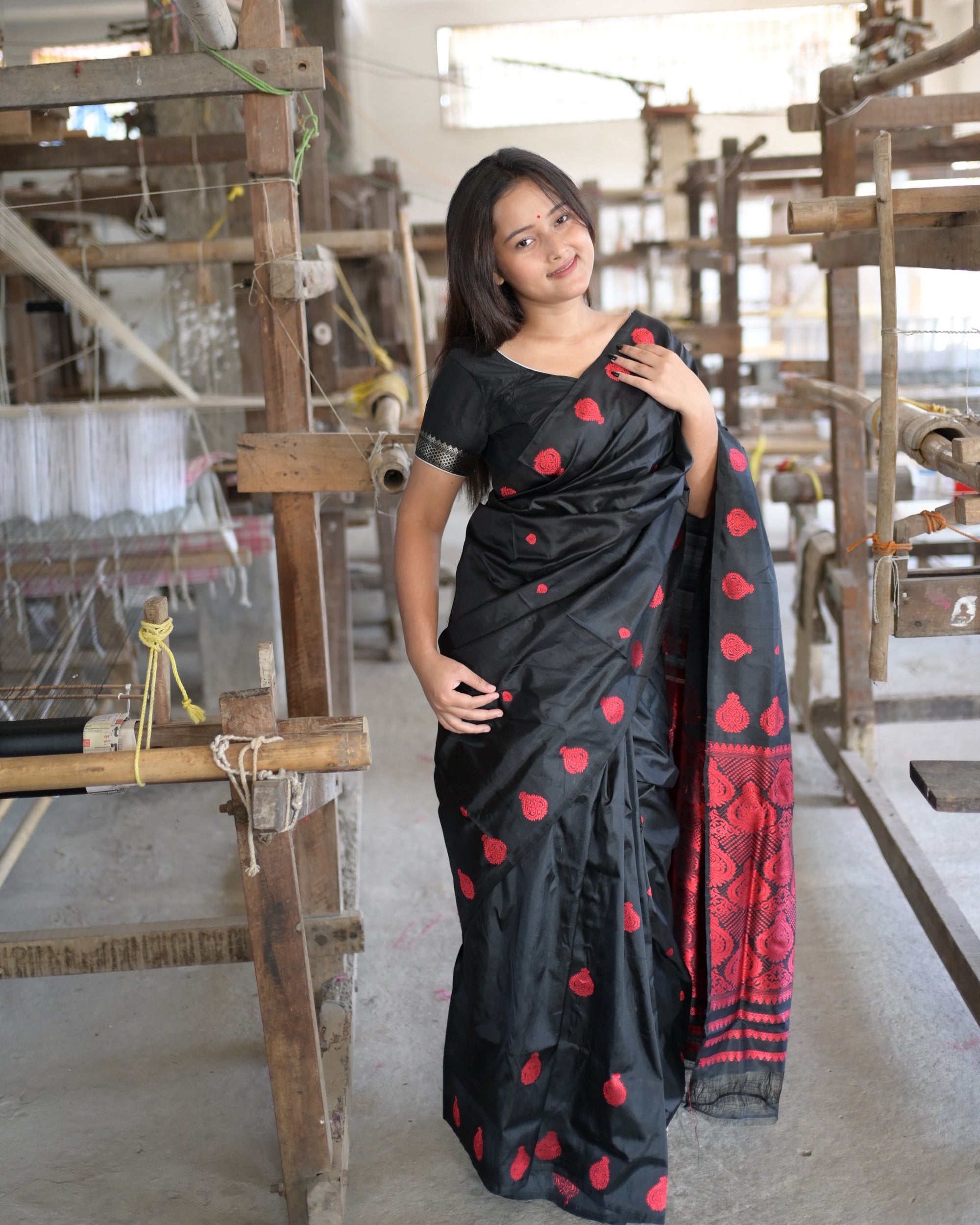 Beside a loom, a hand at the shoulder: red drops scattered over the black body, and the pallu falling over the arm in red and black.
