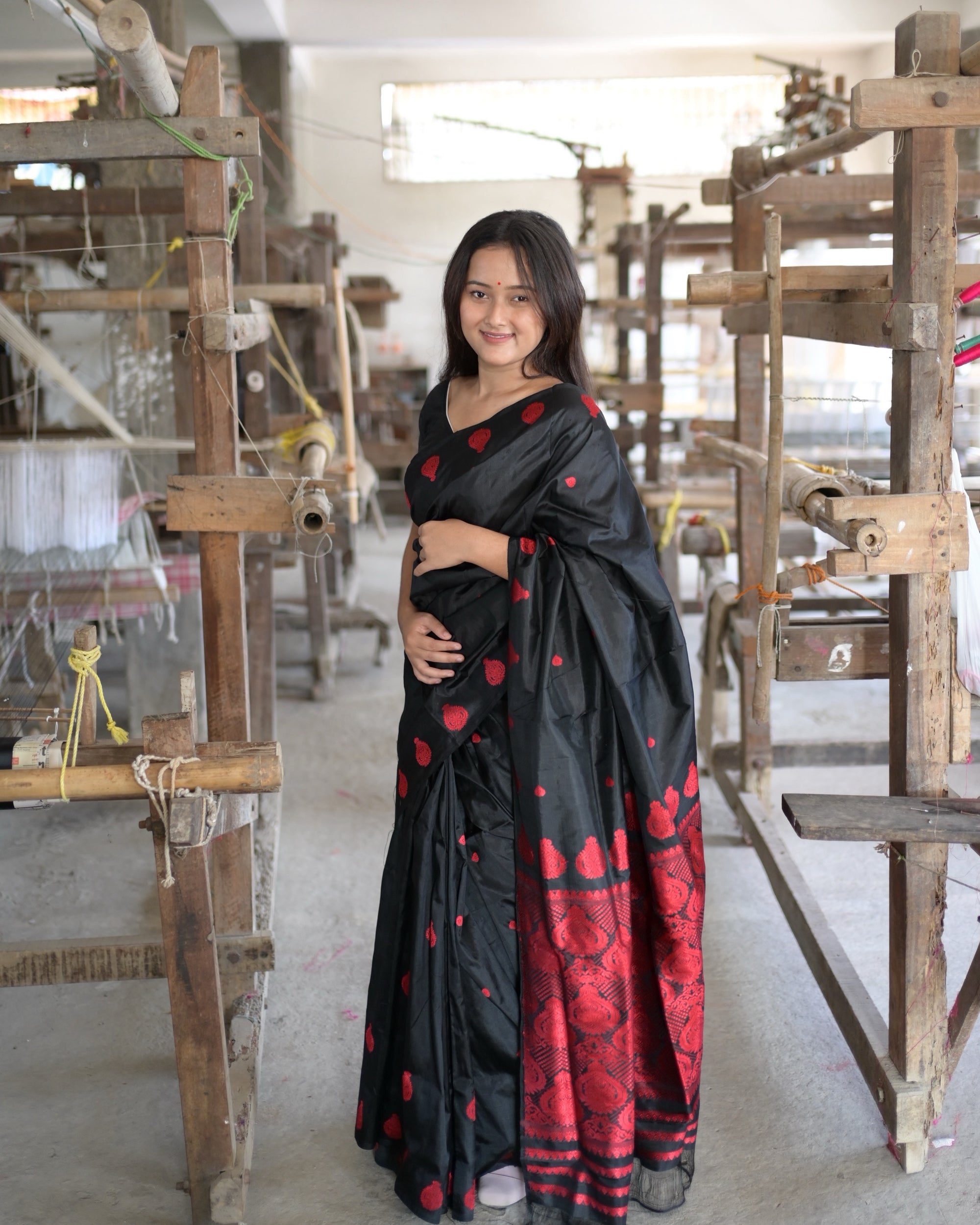 Standing among the handlooms, a hand at the waist: the black body with red drops and a deep red band of medallions along the lower pleats.