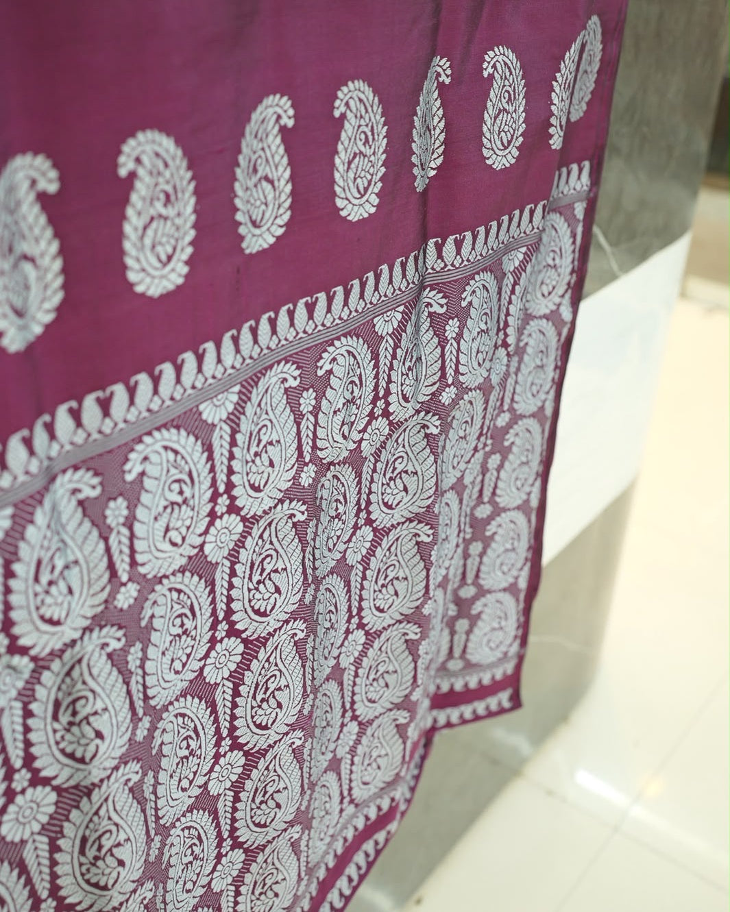 The pallu hanging from the counter, seen at an angle: the row of large paisleys, the curled band and the dense field of paisleys falling towards a pale tiled floor.