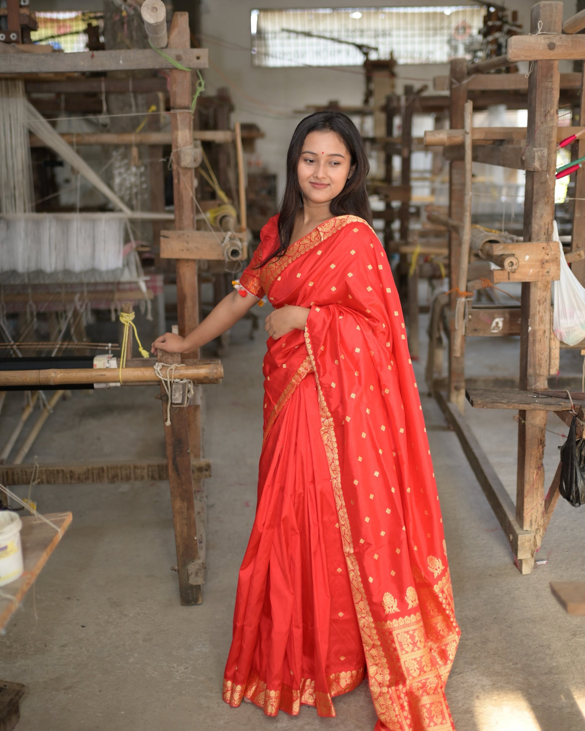 Standing at a handloom in a weaving hall, one hand on its beam, smiling: the bright red saree full length, small golden squares across the body and the broad golden border falling to the floor.