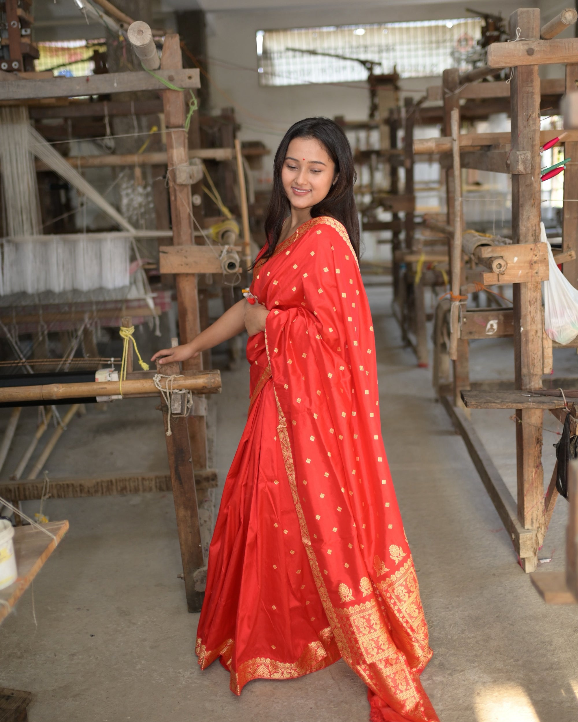 Full length beside a handloom, eyes lowered, one hand on the beam: the red pleats edged in gold and the golden pallu at the hem.