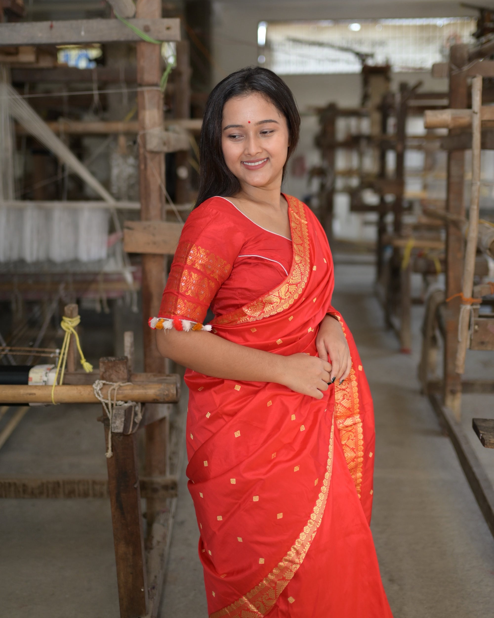 Waist up beside a handloom, eyes lowered and smiling: golden squares on the red silk, the golden border across the shoulder and a red blouse banded in gold at the sleeve.